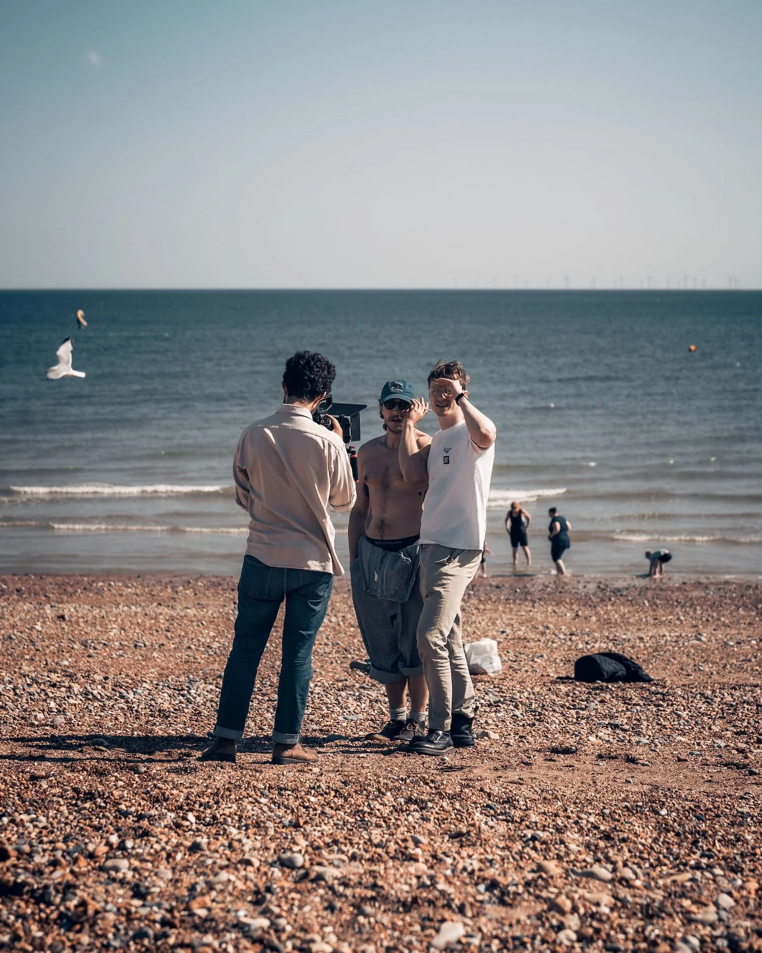 Three people standing on a pebbled beach, with a man and woman being photographed while another man makes a gesture and smiles. In the background, there are people near the water, seagulls flying, and an ocean horizon.