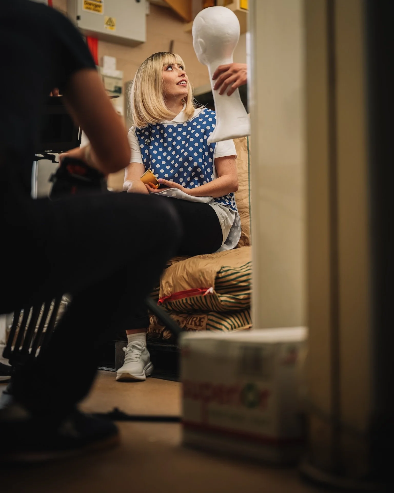A woman with blonde hair and a white and blue polka dot shirt sitting on a bed in a room, looking up at a person holding a mannequin head. The photo is taken from a low angle through some objects in the foreground.