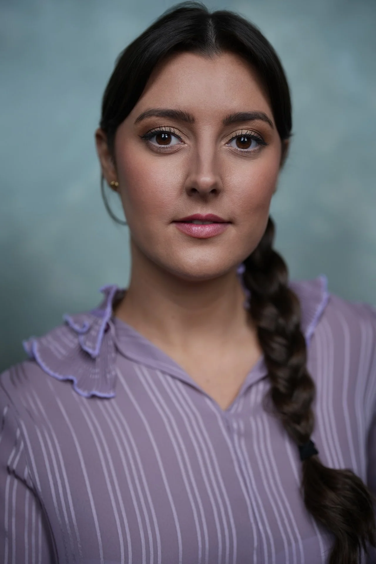A close-up portrait of a woman with dark brown hair in a braid, wearing a purple striped blouse, with a blurred grey-green background.