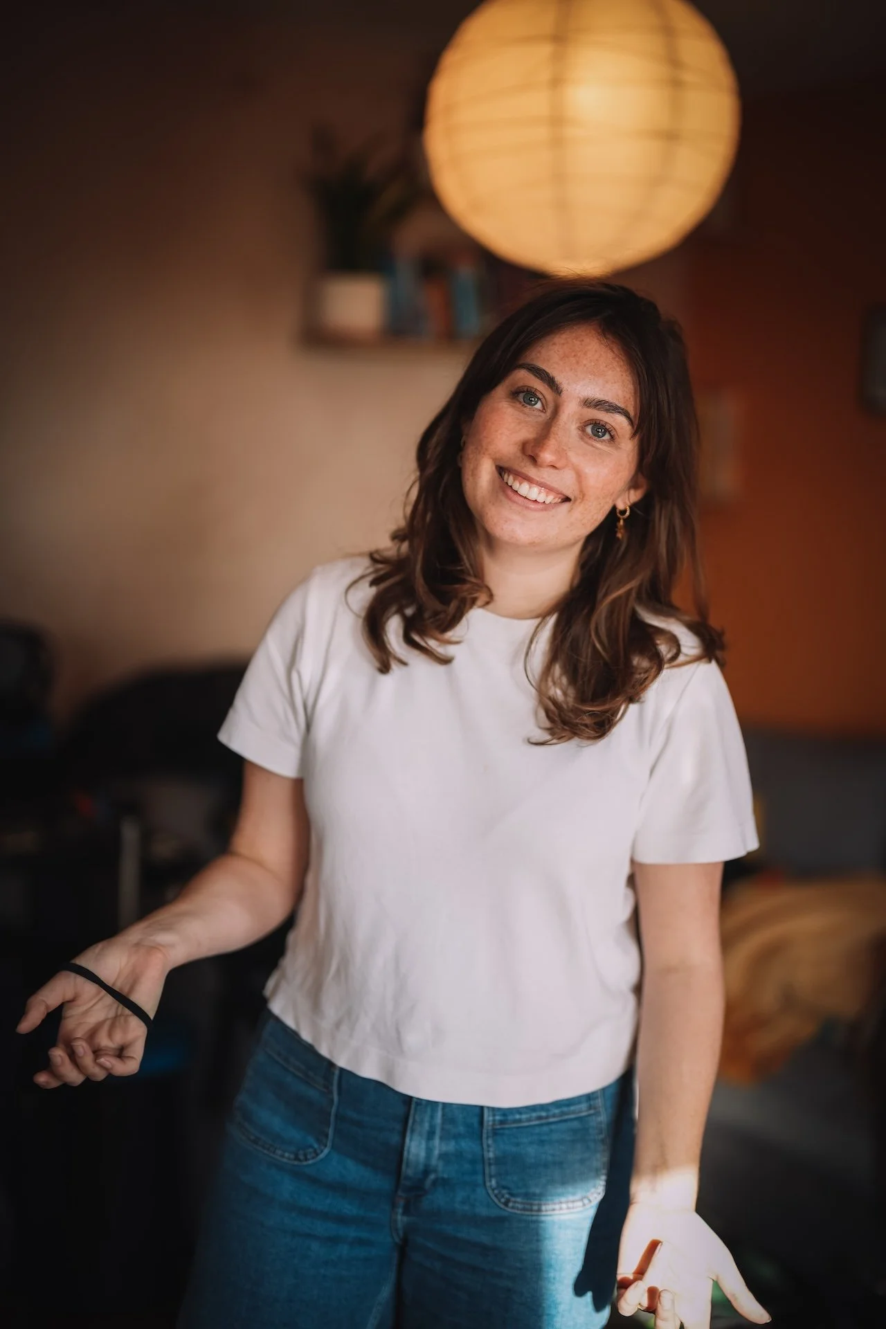 A smiling woman with brown hair and freckles wearing a white t-shirt and jeans in a warmly lit room.