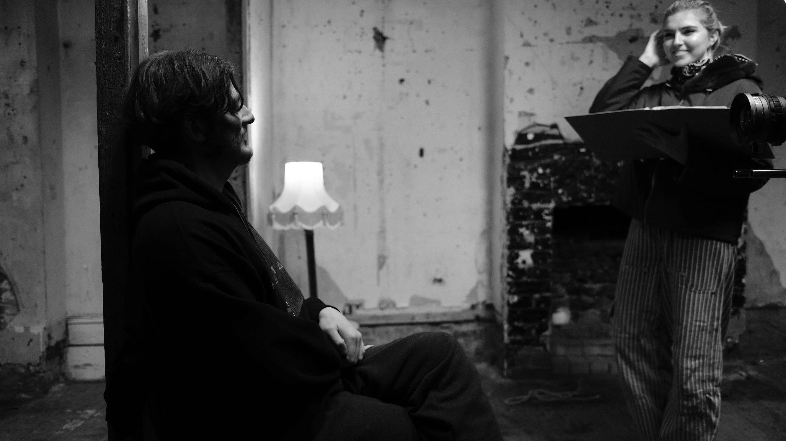 A man sitting against a wall in conversation with a woman standing nearby, holding a folder and camera in a dimly lit, rustic room.