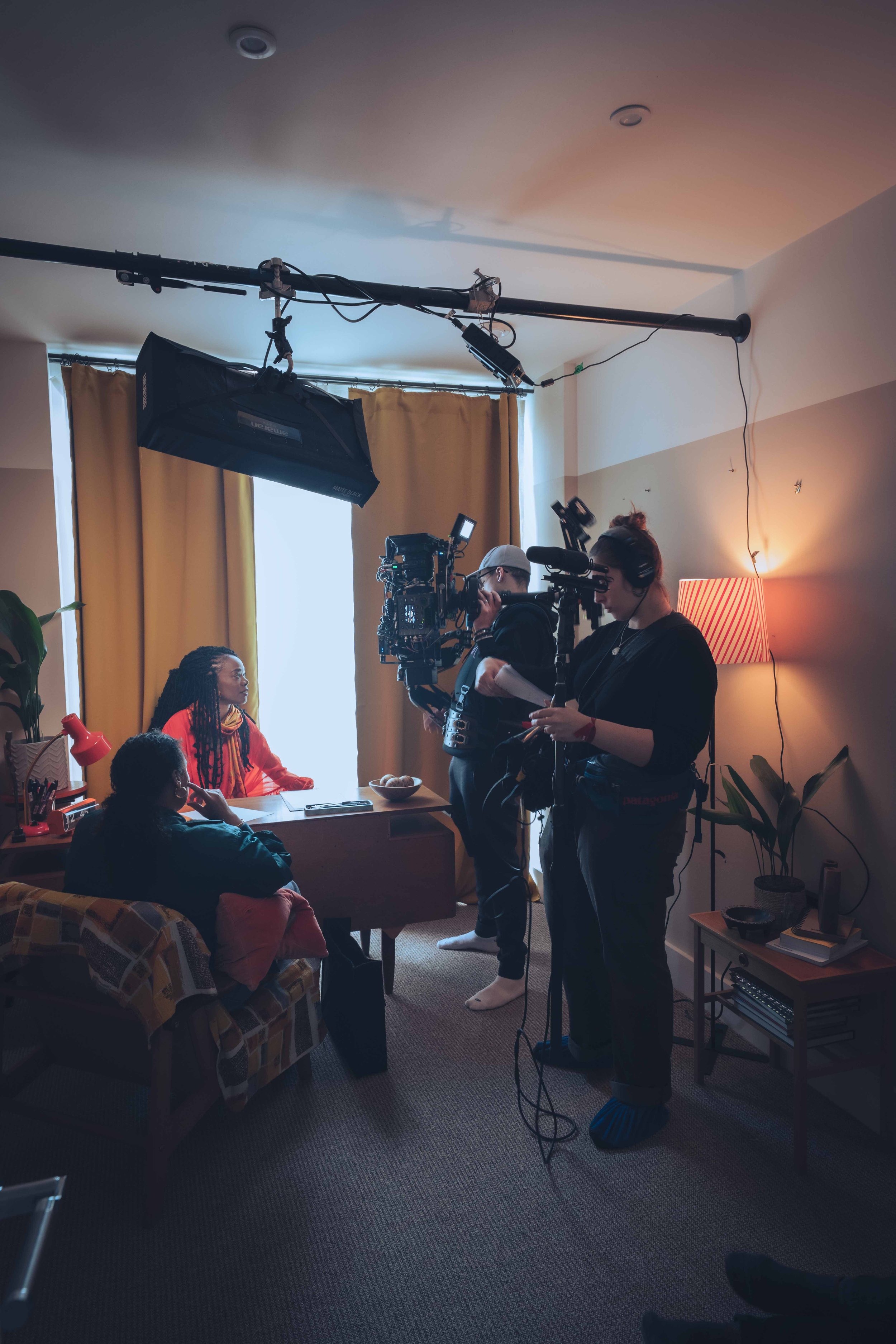 Behind-the-scenes of a video or photo shoot in a cozy living room. Crew members are setting up equipment, including a camera, lights, and a boom microphone. Two women are seated, one with dreadlocks and colorful clothing, and the other wearing glasse