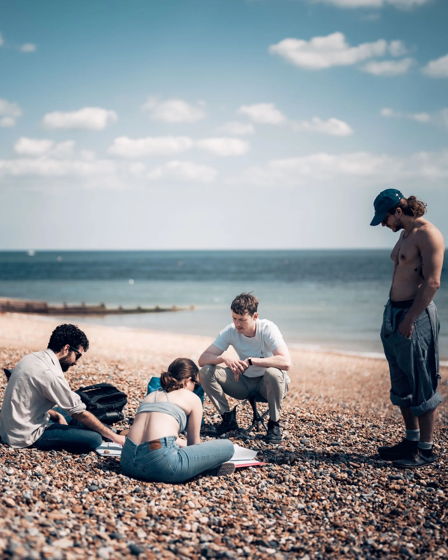 A group of people on a rocky beach, with some sitting and others standing, gathered around a woman who is lying on the ground. The background shows the ocean and a partly cloudy sky.