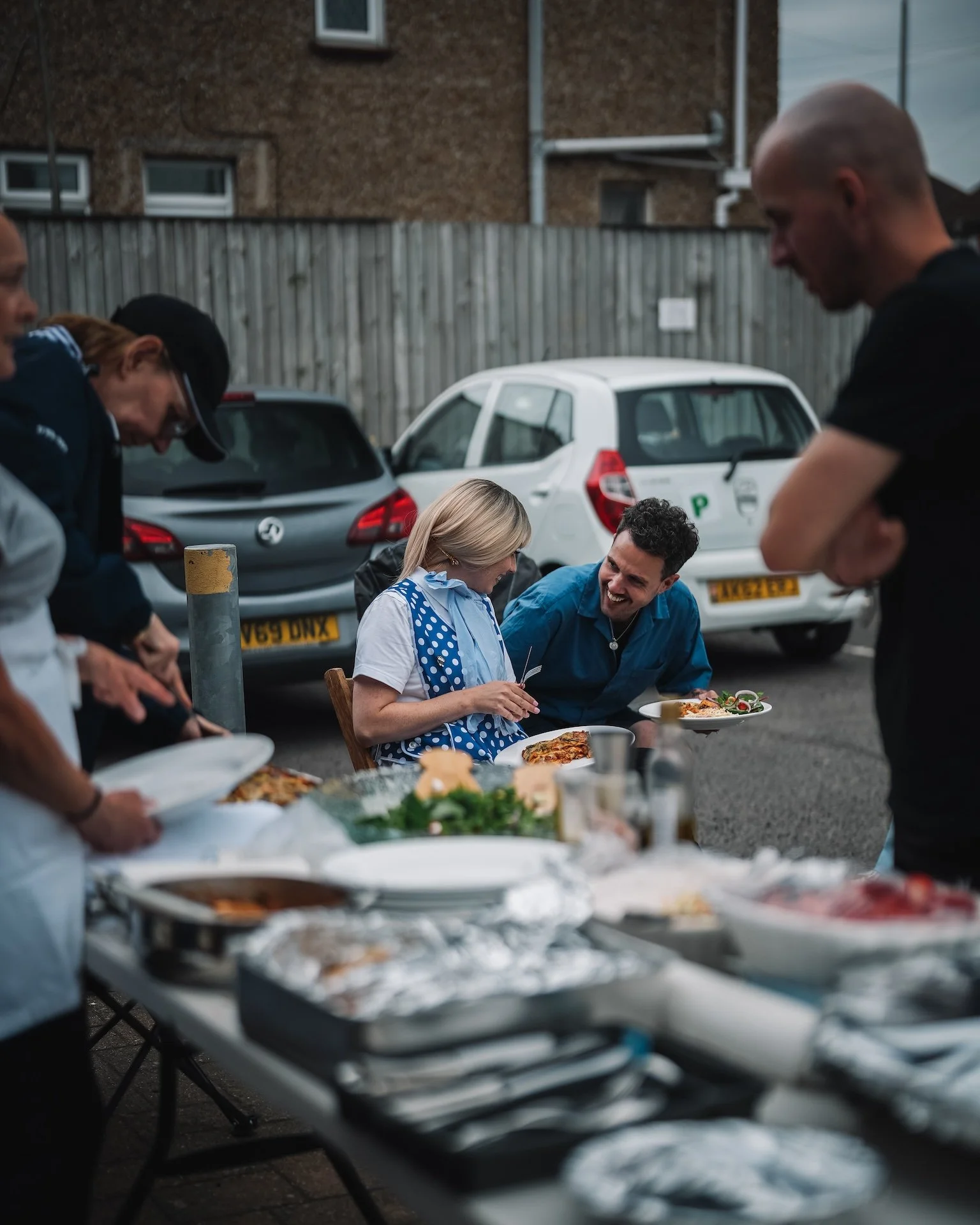 People gathered outdoors for a meal, with tables full of food, pizza, and drinks, in an urban setting near parked cars and a brick building.