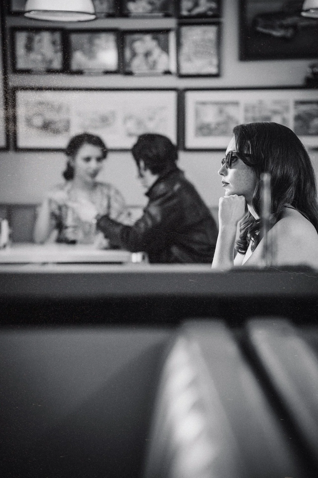 A woman with dark hair and glasses sitting at a table in a restaurant, resting her chin on her hand. In the background, two people are talking and there are various framed pictures on the wall.