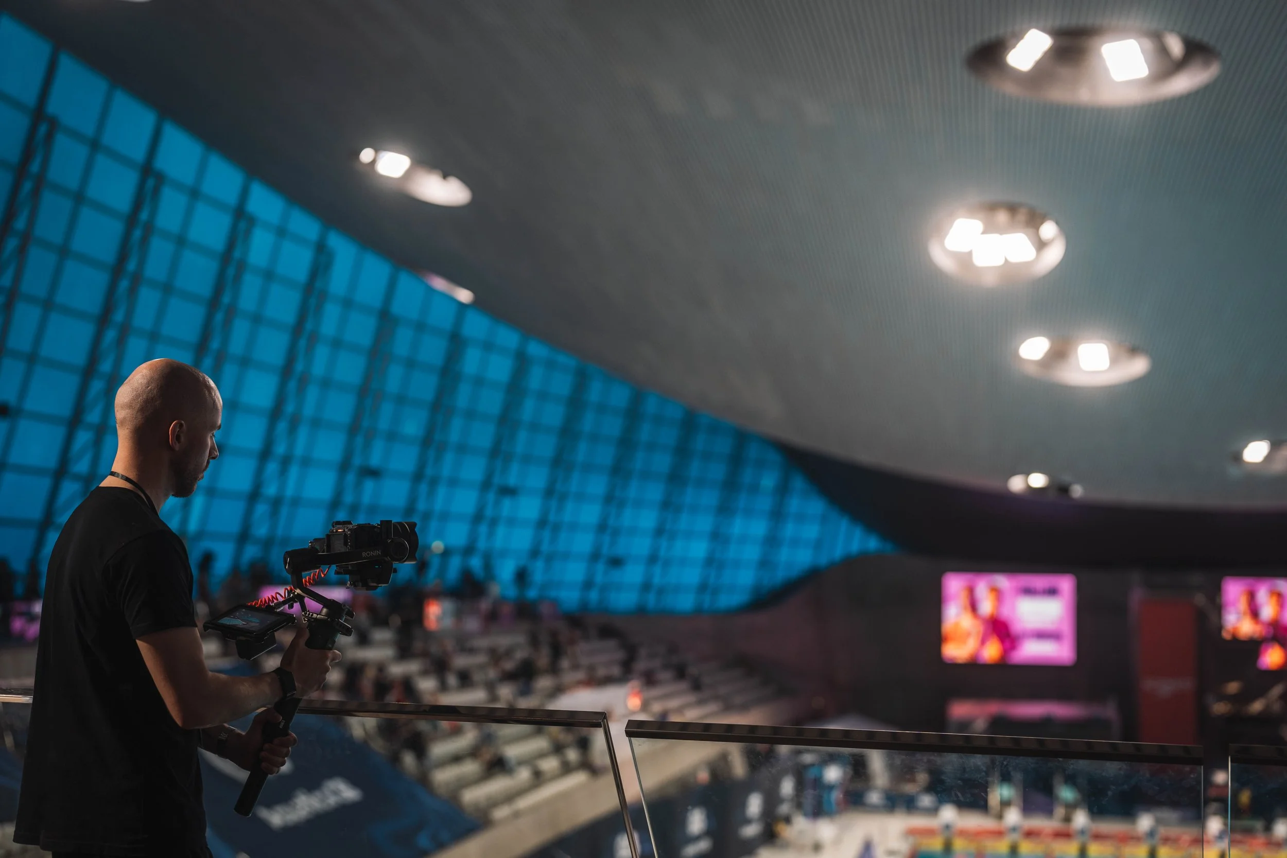A man operating a camera on a stabilizer in a large indoor stadium with high, curved ceiling and bright ceiling lights.