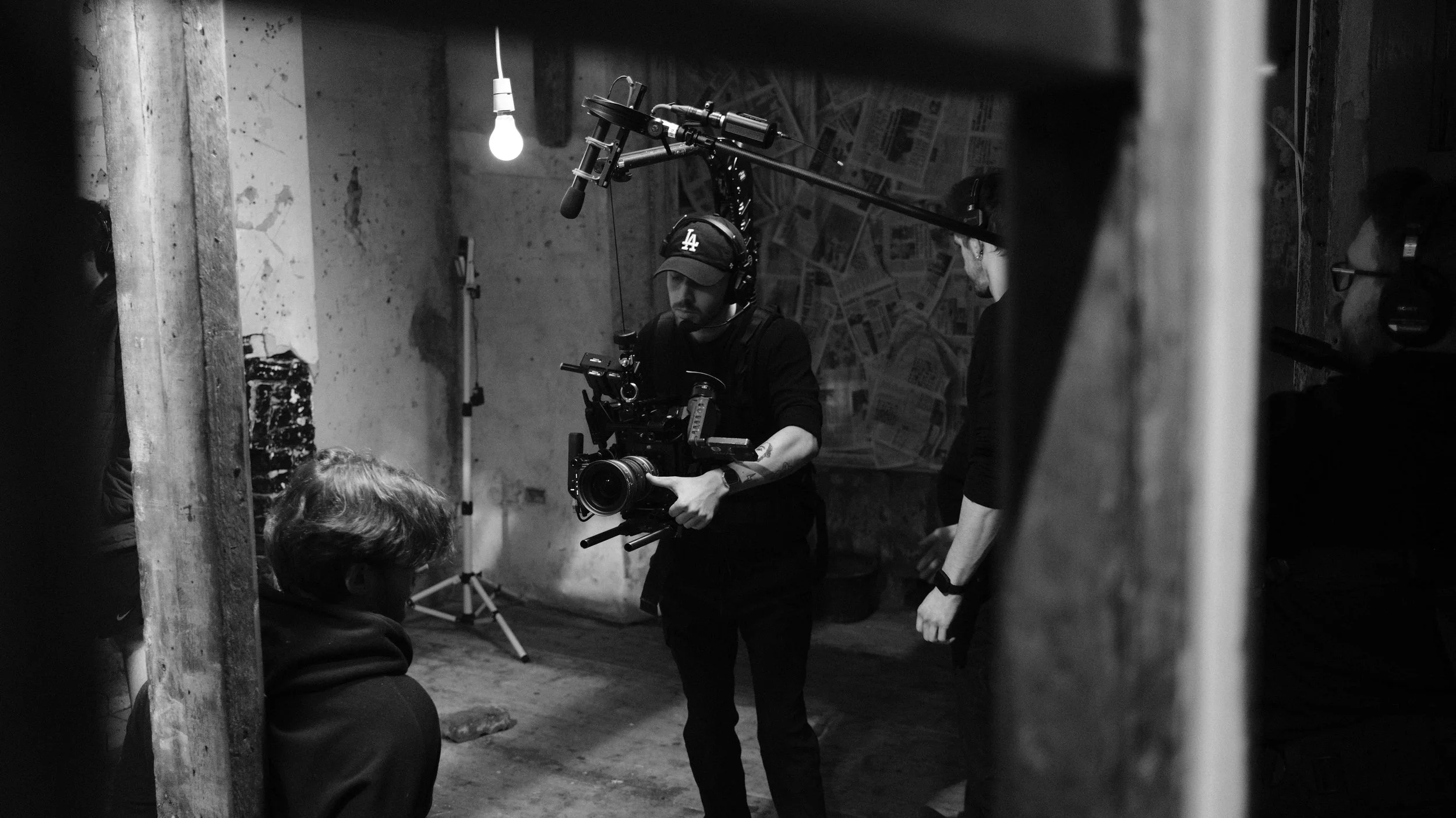 A black and white photo of a film crew shooting a scene, with a camera operator holding a professional camera and a crew member holding a boom microphone, in a dimly lit, rough indoor setting.