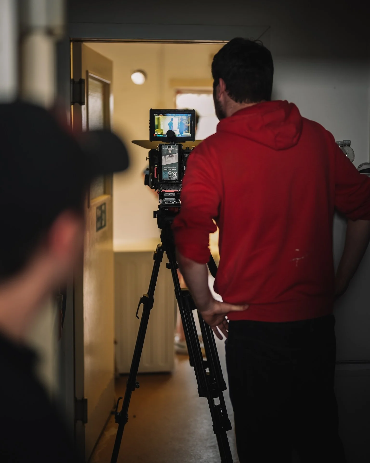 A man in a red hoodie operating a camera on a tripod, filming a scene with a bright window and two figures on a monitor screen.