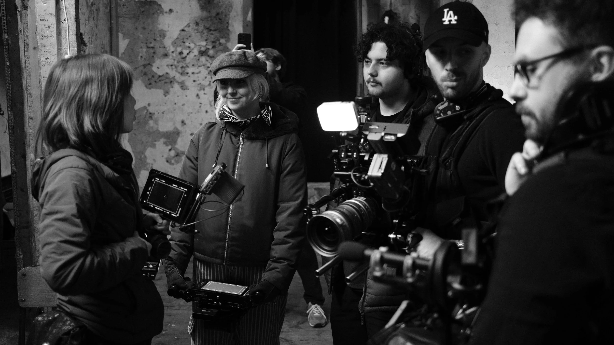 A group of five people, including a woman with a camera, engaged in a filming or photography session in an urban space with distressed walls. The scene is in black and white.
