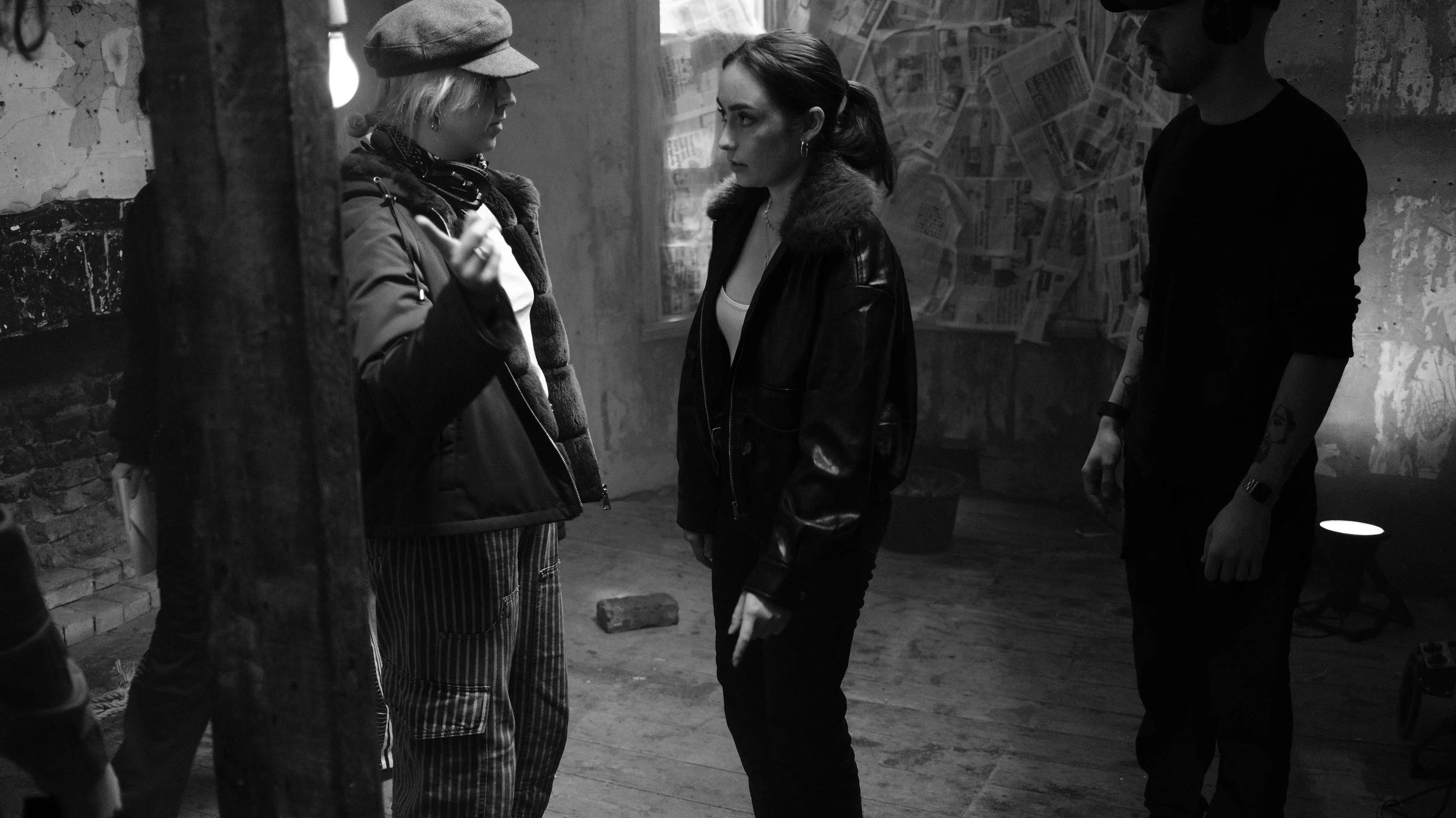 Three people standing in a dimly lit, grungy room with newspaper-covered windows and exposed brick walls, engaging in conversation.