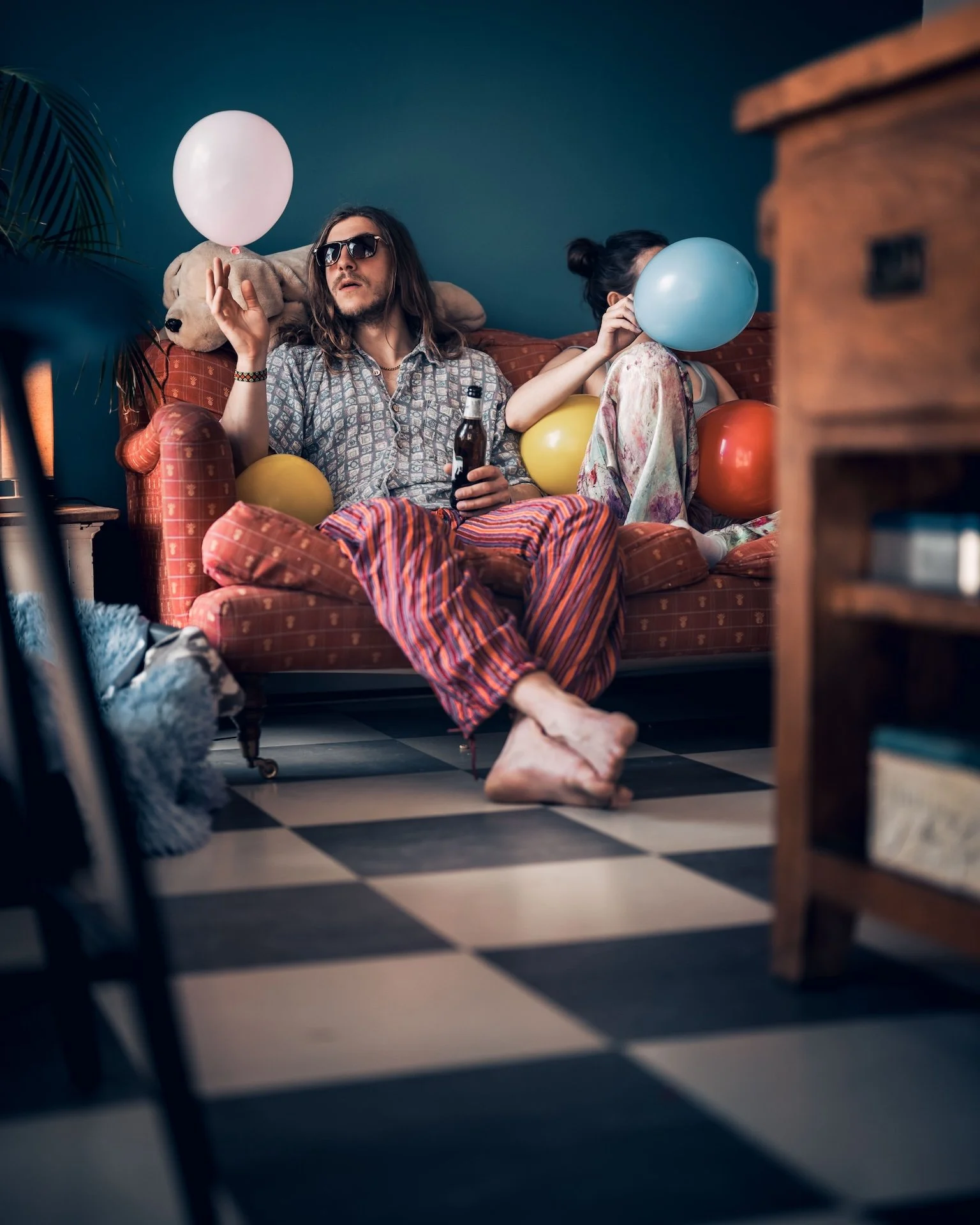 A man with long hair and sunglasses sitting on a red sofa with patterned fabric, wearing striped pajama pants, holding a beer bottle, with a woman beside him holding balloons, in a room with a checkered black and white floor, a wooden furniture piece