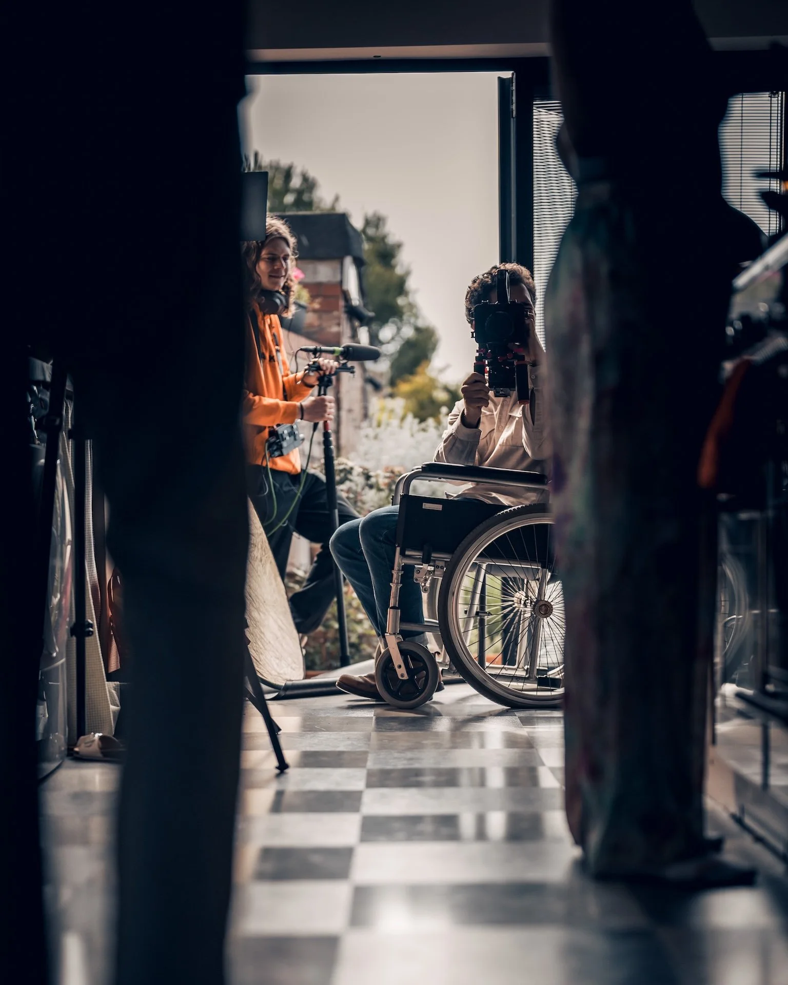 Inside view of a house, looking through a narrow gap between people and objects, showing a woman with headphones and a man in a wheelchair with a camera, with outdoor scenery in the background.