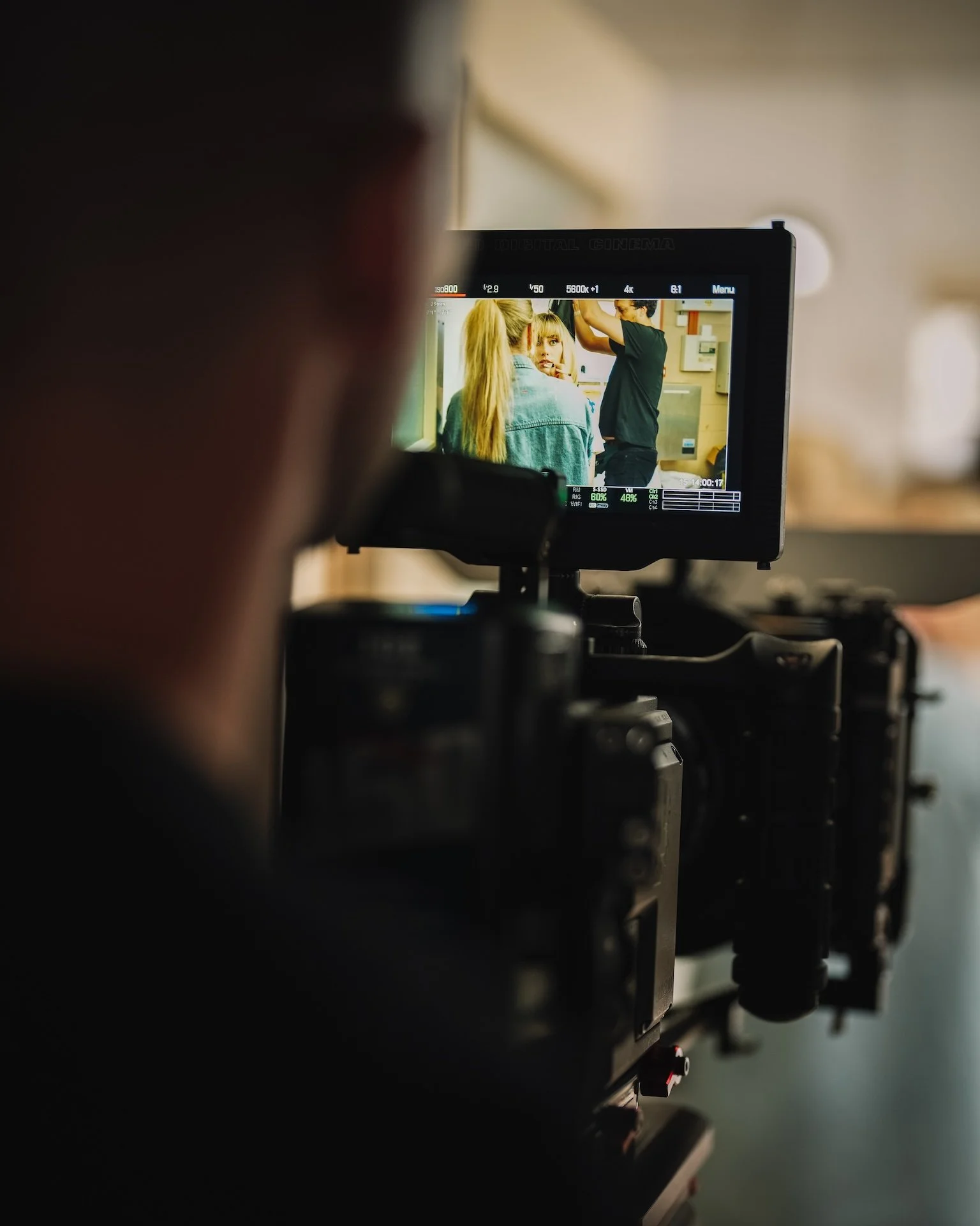 Behind-the-scenes view of a film or video shoot, with a monitor showing two women, one with blonde hair and a denim jacket, and a man adjusting her hair, in a room.
