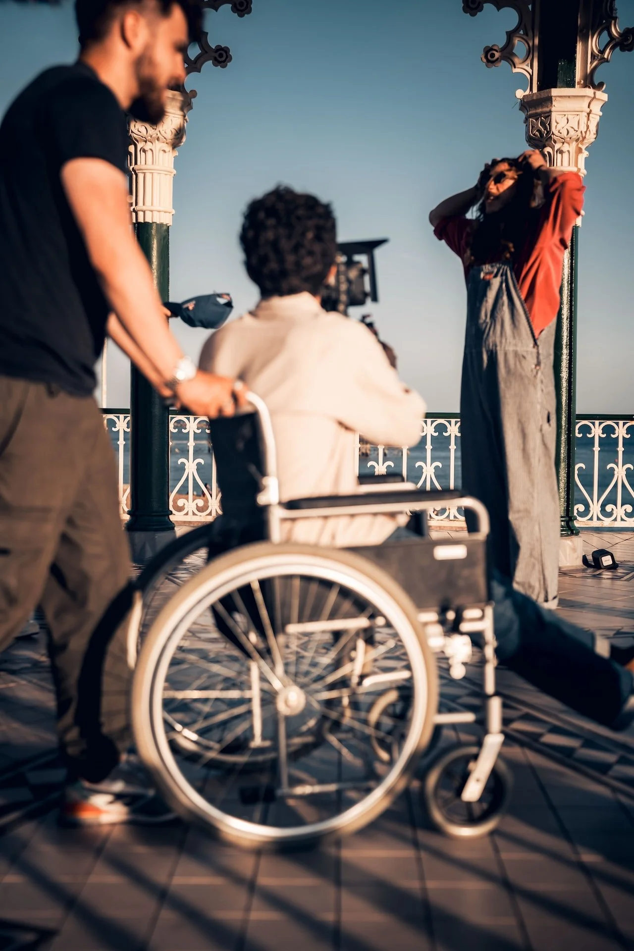 A person in a wheelchair on a patterned floor, with two other people nearby, outdoors on a terrace or balcony with ornate columns and a railing, during sunset or golden hour.