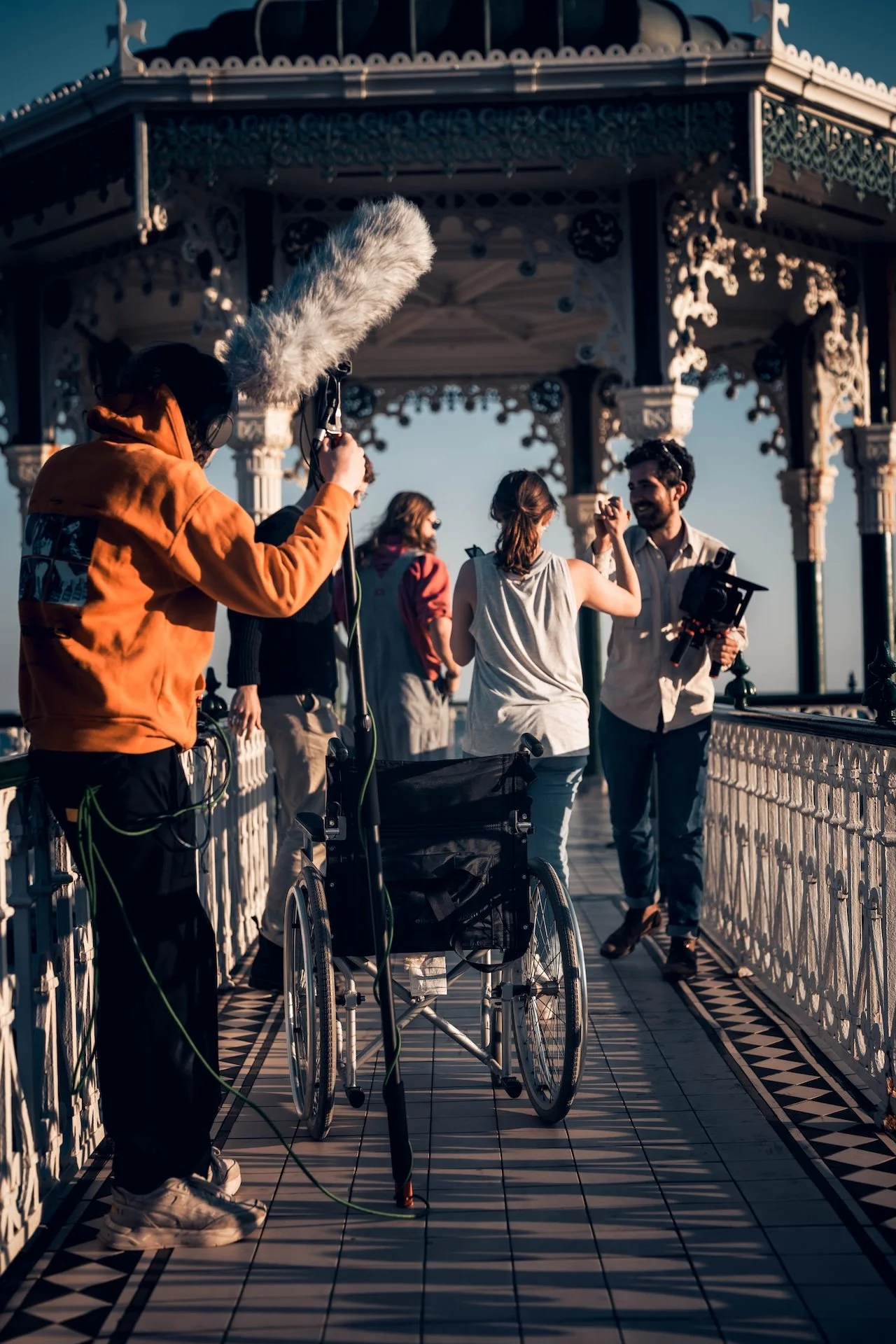 Filmmakers filming outdoors under a decorative gazebo, with three women and a man, one woman in a wheelchair, on a patterned tile walkway.