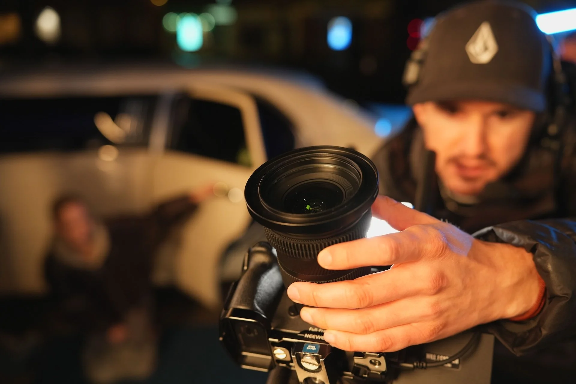 A person wearing a helmet operating a drone with a camera, holding a remote control at night with blurred city lights in the background.