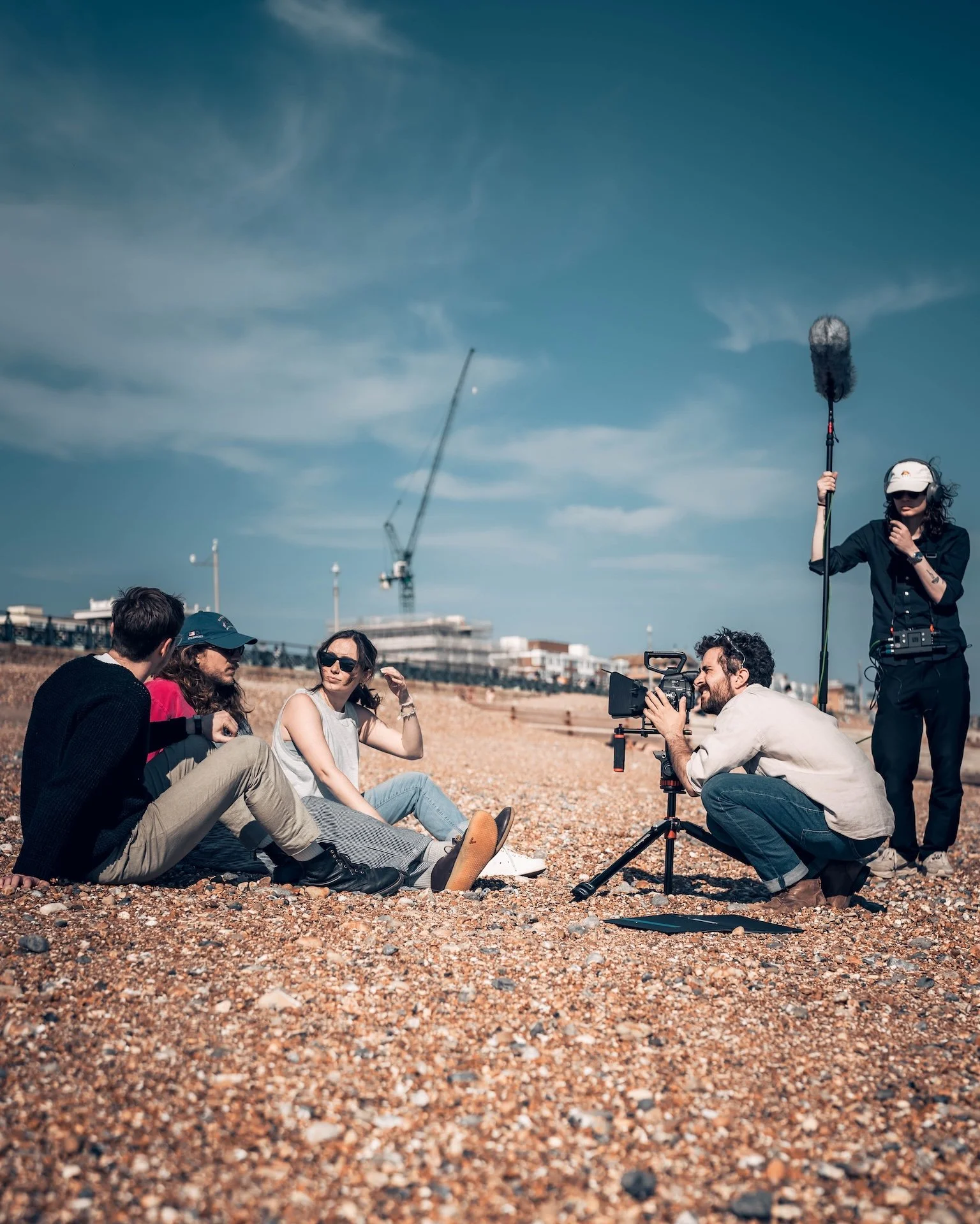 Group of people sitting on a rocky outdoor area during a photoshoot, with a woman operating a microphone, a cameraman capturing the scene, and several individuals engaged in conversation against a background of blue sky and construction cranes.
