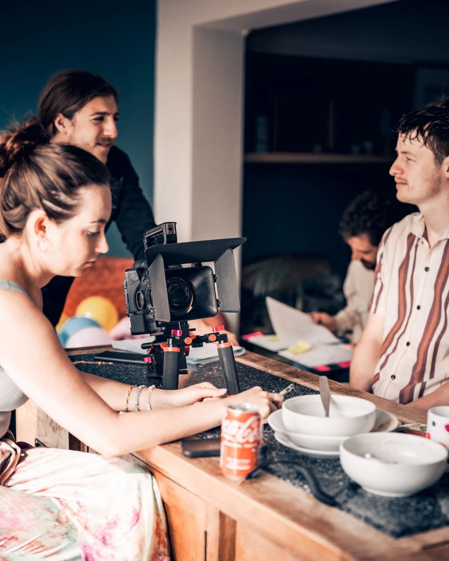 People sitting at a kitchen table, with one person operating a camera on a tripod, food, drinks, and food containers on the table.