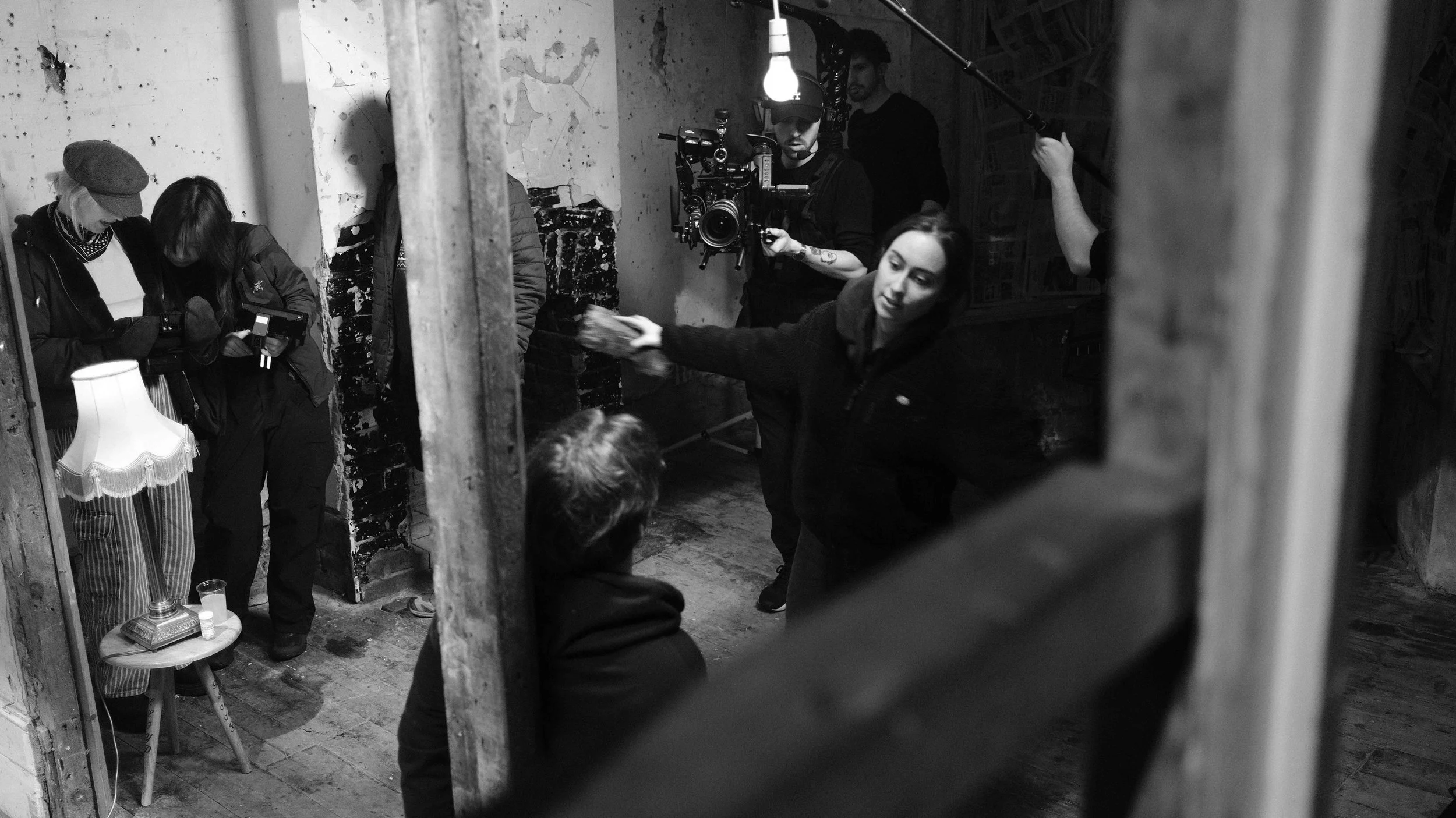Film crew working on a set with a director giving instructions, a camera operator with a camera rig, and crew members adjusting equipment, in a rustic, industrial space with exposed brick and distressed walls.