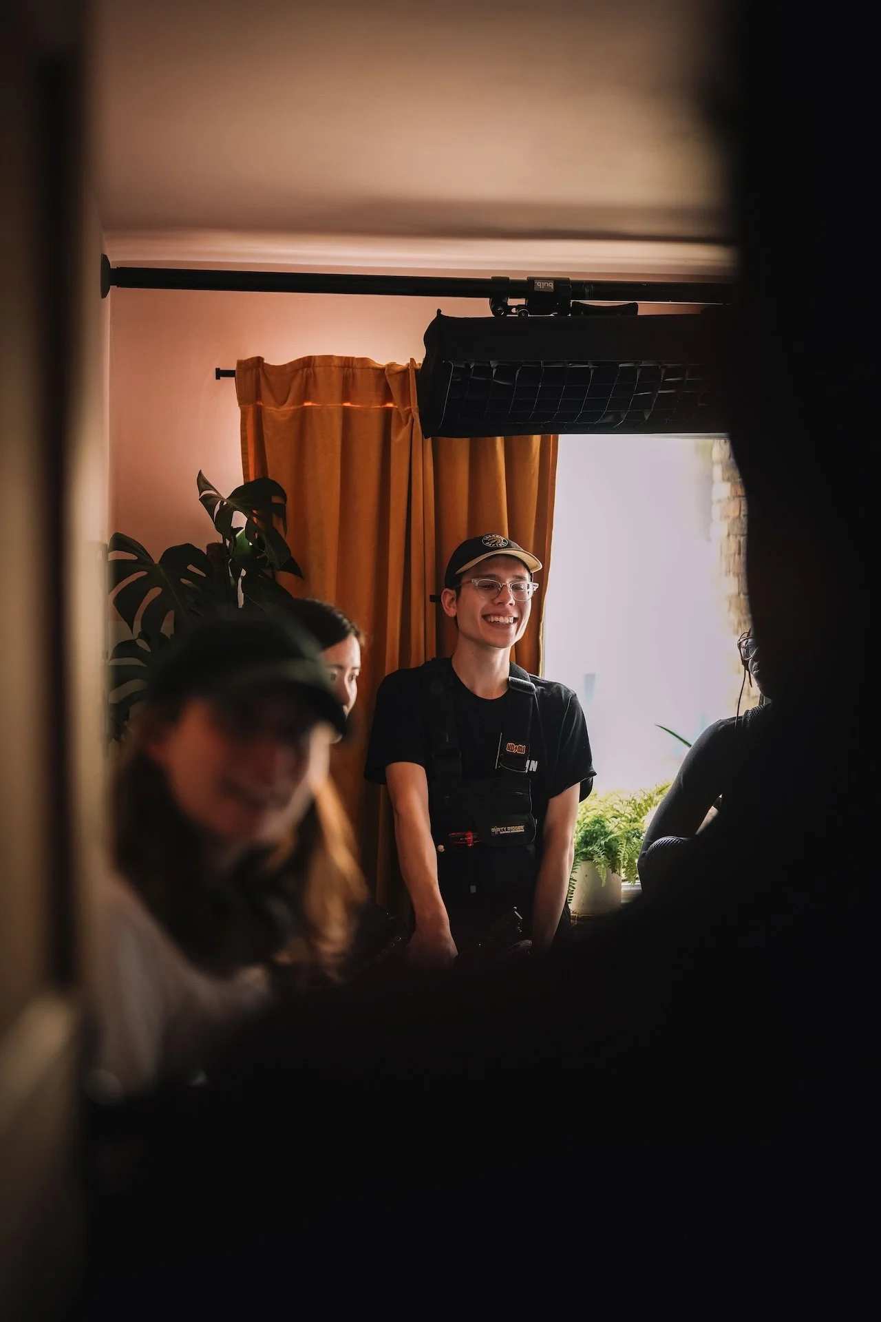 Young man wearing glasses and a baseball cap smiling, standing by a window with orange curtains inside a room with people.