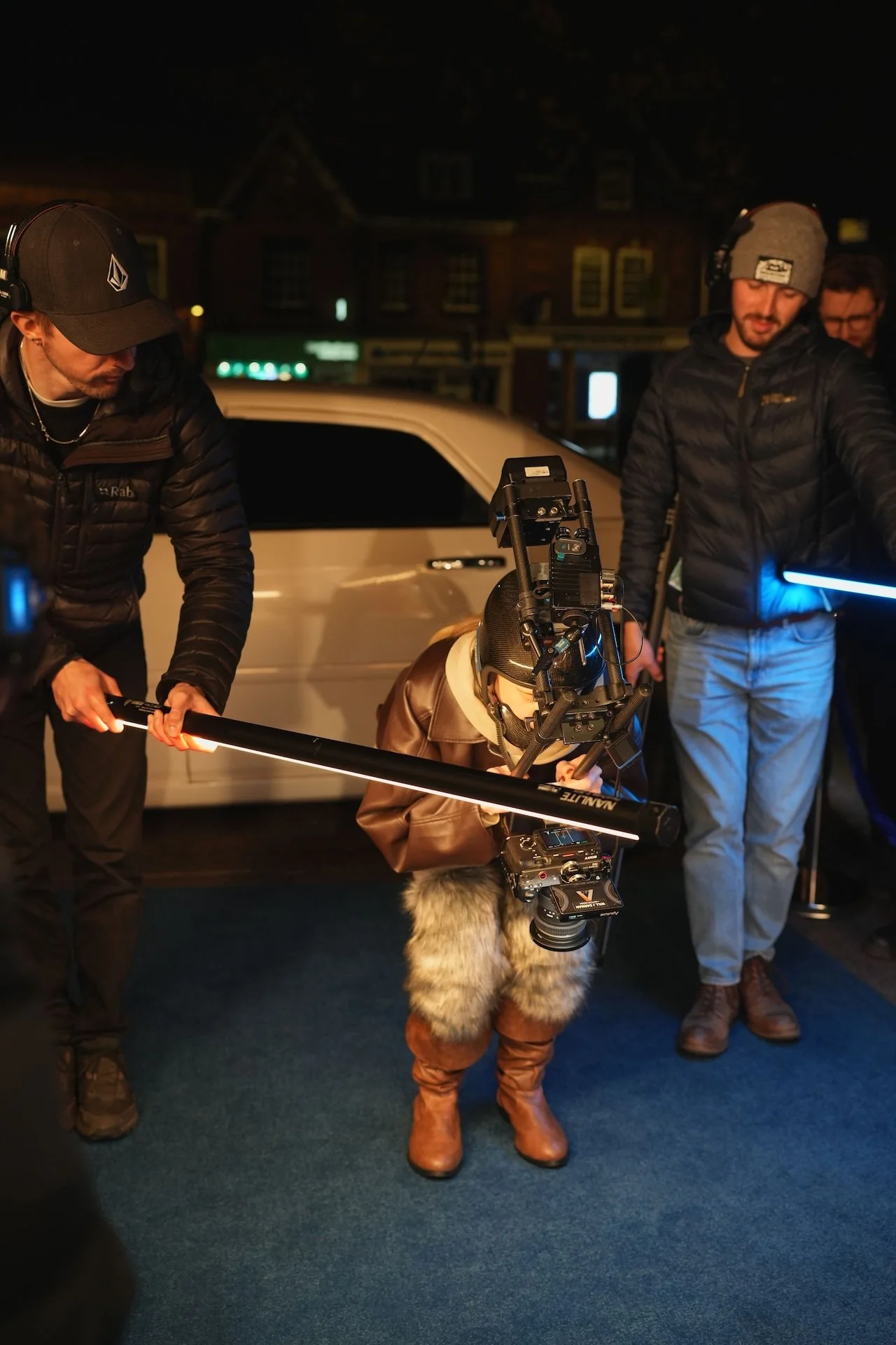 A film crew setting up equipment outdoors at night, with crew members wearing jackets and headphones, a white car in the background, and a person with a camera rig and fur-lined boots adjusting equipment.