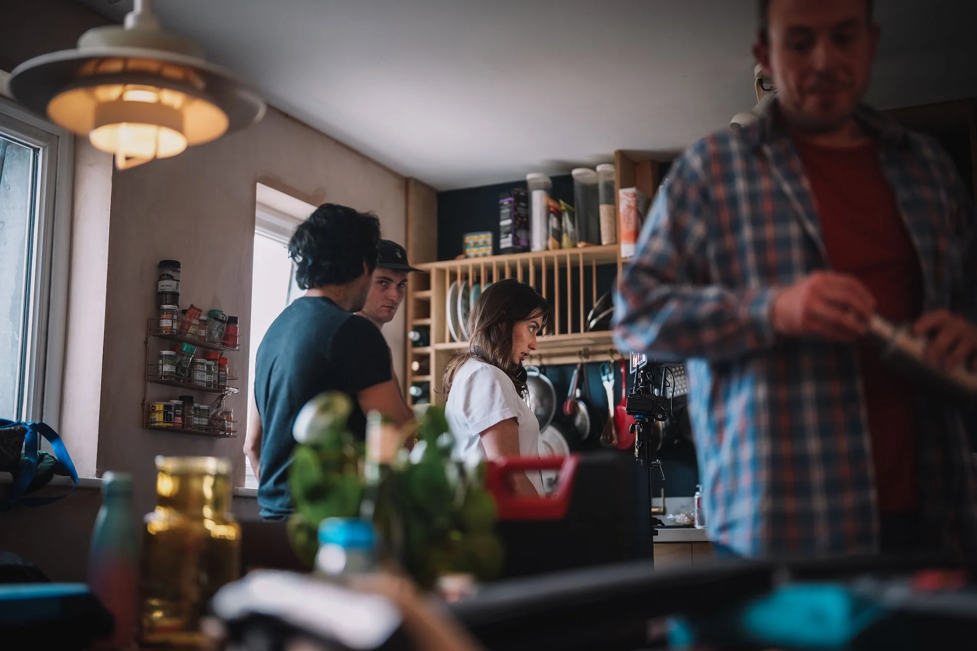 People gather in a kitchen, some engaged in conversation and others focused on tasks, with shelves of dishes and spices visible in the background.