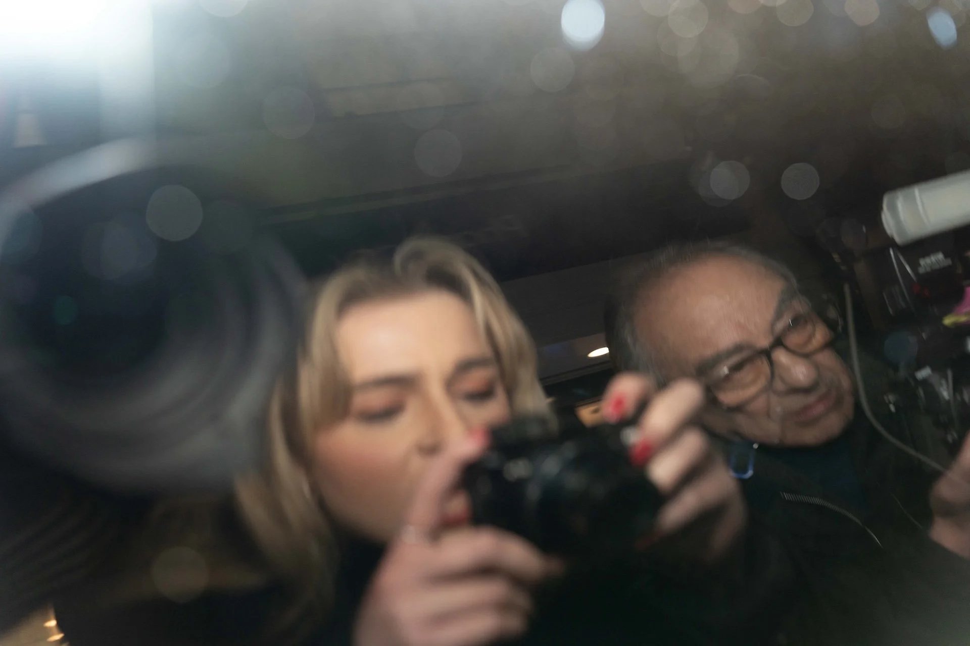 Two people taking a selfie or a photo, with a camera lens partially visible in the foreground. The woman has long blonde hair, and the man has glasses and dark hair; they are smiling and appear to be in a well-lit interior space.