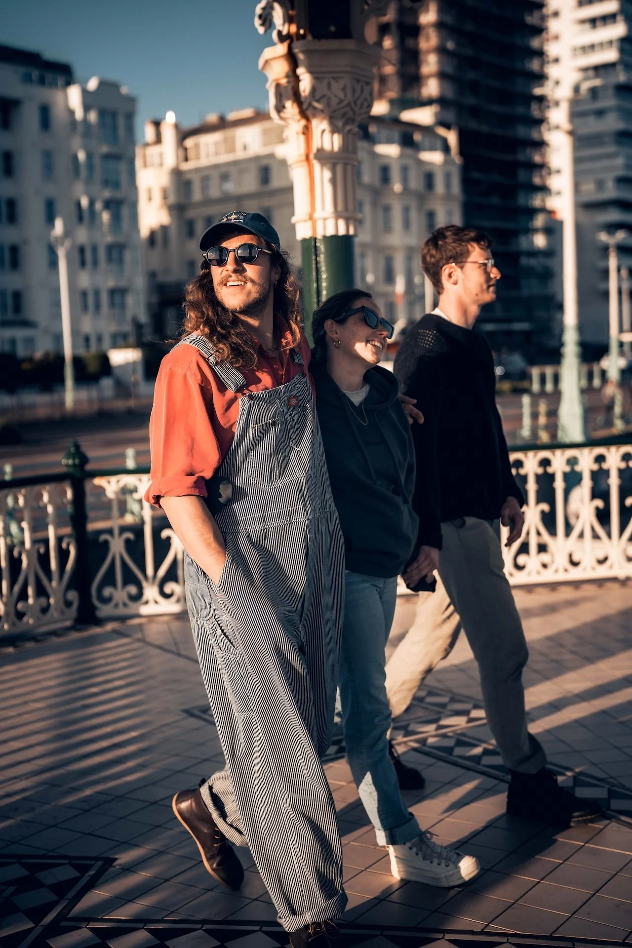 Three friends walking outdoors in a city, enjoying sunny weather, with tall buildings in the background. The person in the front is wearing overalls and sunglasses, smiling. The others are walking alongside, also wearing sunglasses and casual clothes