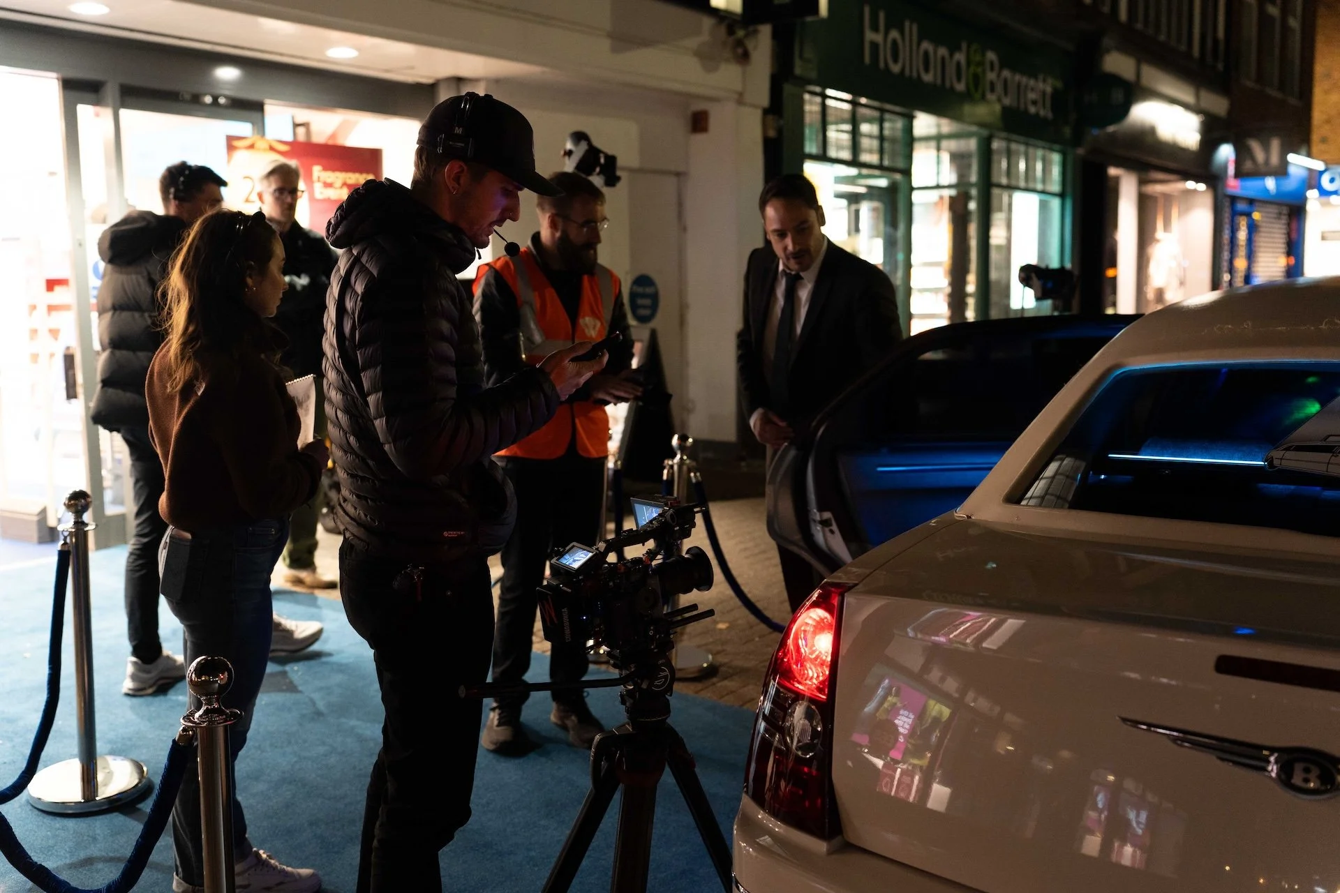 Filmmakers and crew filming a scene at night outside a car dealership, with a camera mounted on a stabilizer and crew members observing and managing the equipment.