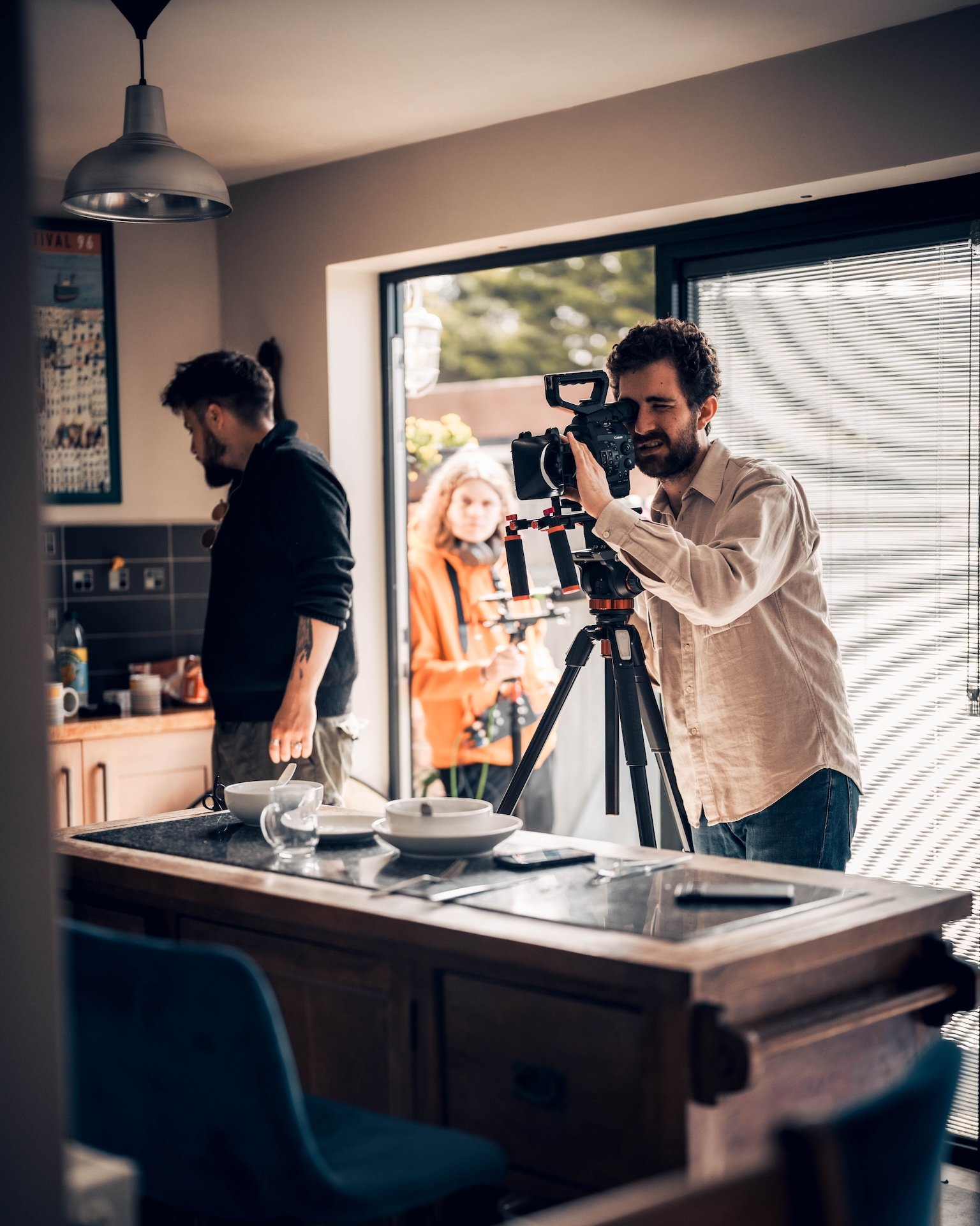 A man filming with a professional camera in a modern kitchen, with another person preparing food in the background, and a woman outside visible through the glass door.