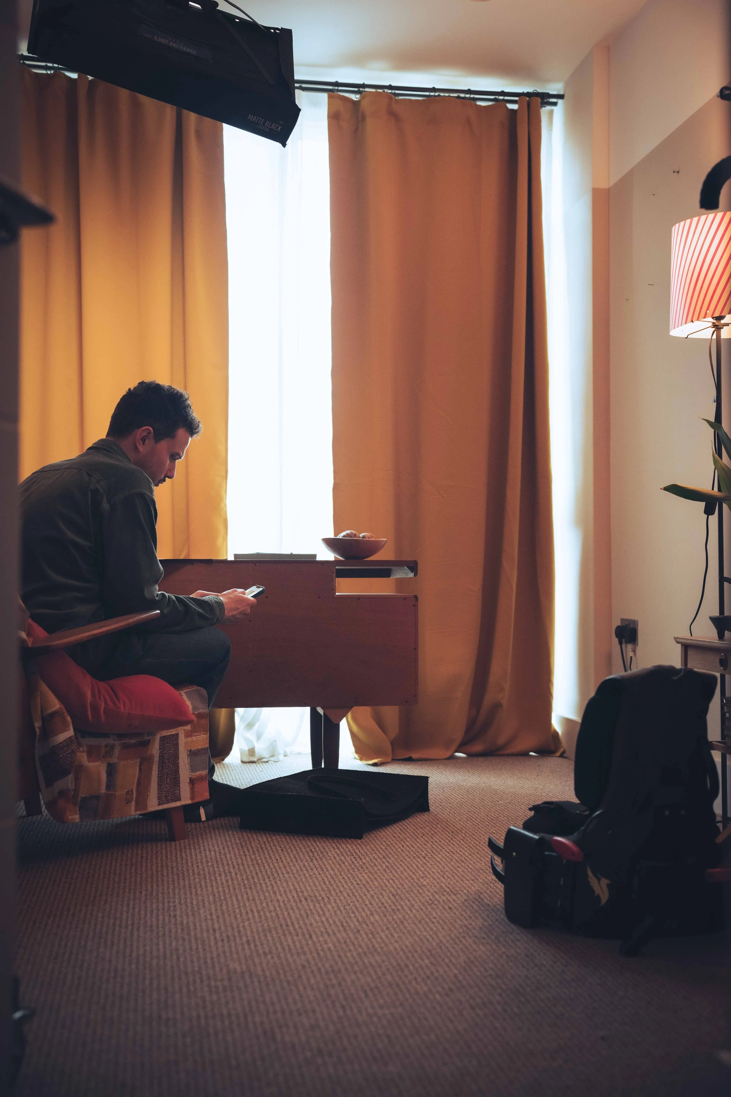 Man sitting on a chair using a smartphone in a room with yellow curtains, a lamp with a red and white striped shade, and camera equipment, including a softbox and a bag on the floor.