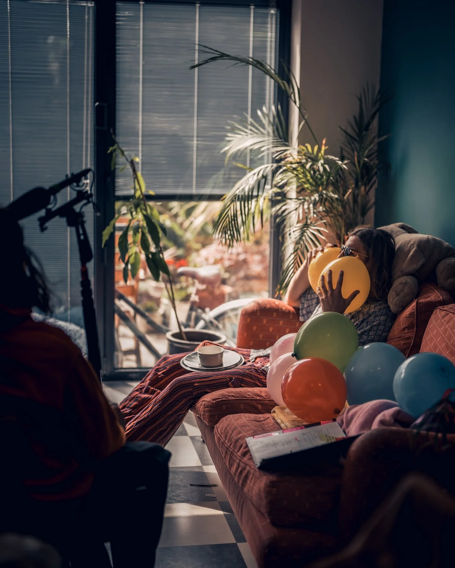 A woman with glasses blows up balloons for a celebration in a living room, with balloons on a table and a person's arm in the foreground, near large plants and a window with blinds.