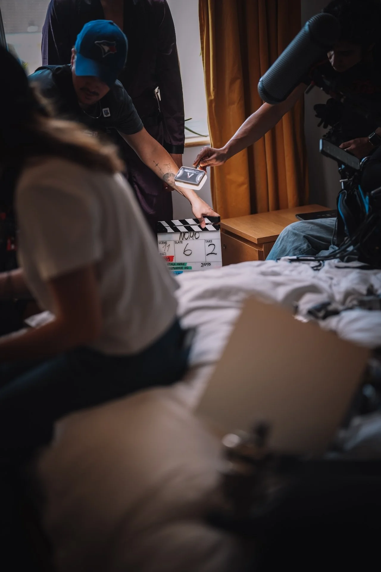 On a film set, crew members are filming a scene in a bedroom while a person lies on a bed. The crew uses a clapperboard with scene and take details, and one person holds a clipboard, possibly with notes or a script. The room has a window with curtain