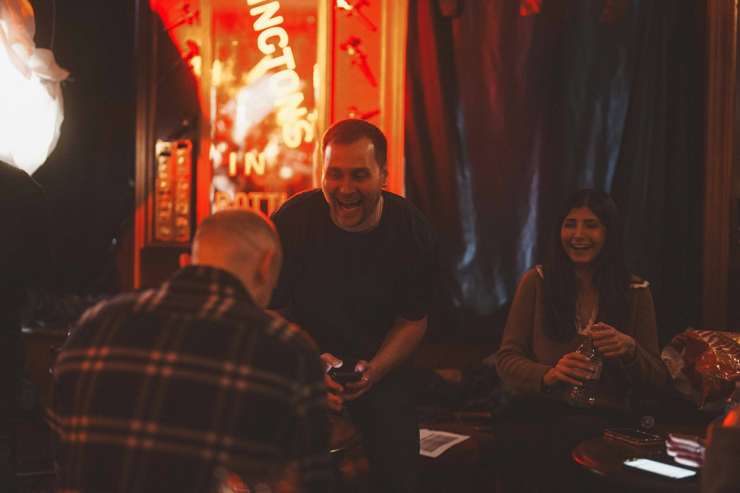 People gathered in a dimly lit setting, possibly a bar or restaurant, with warm red and orange lighting. A man is laughing and holding a mobile phone, while others sit around him, engaging in conversation. A neon sign and dark curtains are visible in