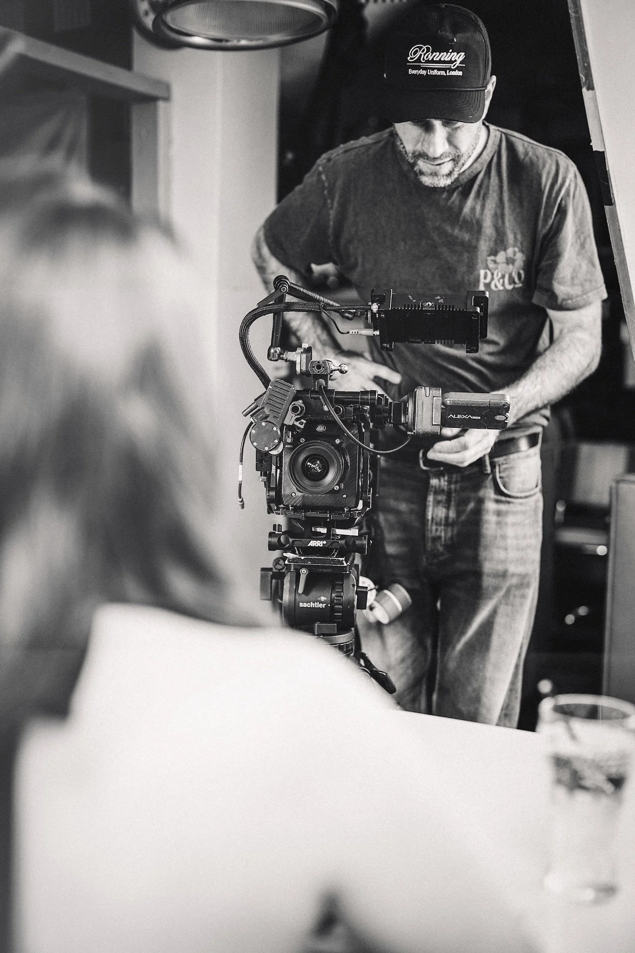 A man wearing a baseball cap and casual shirt operates a professional camera on a tripod, focusing on filming indoors.