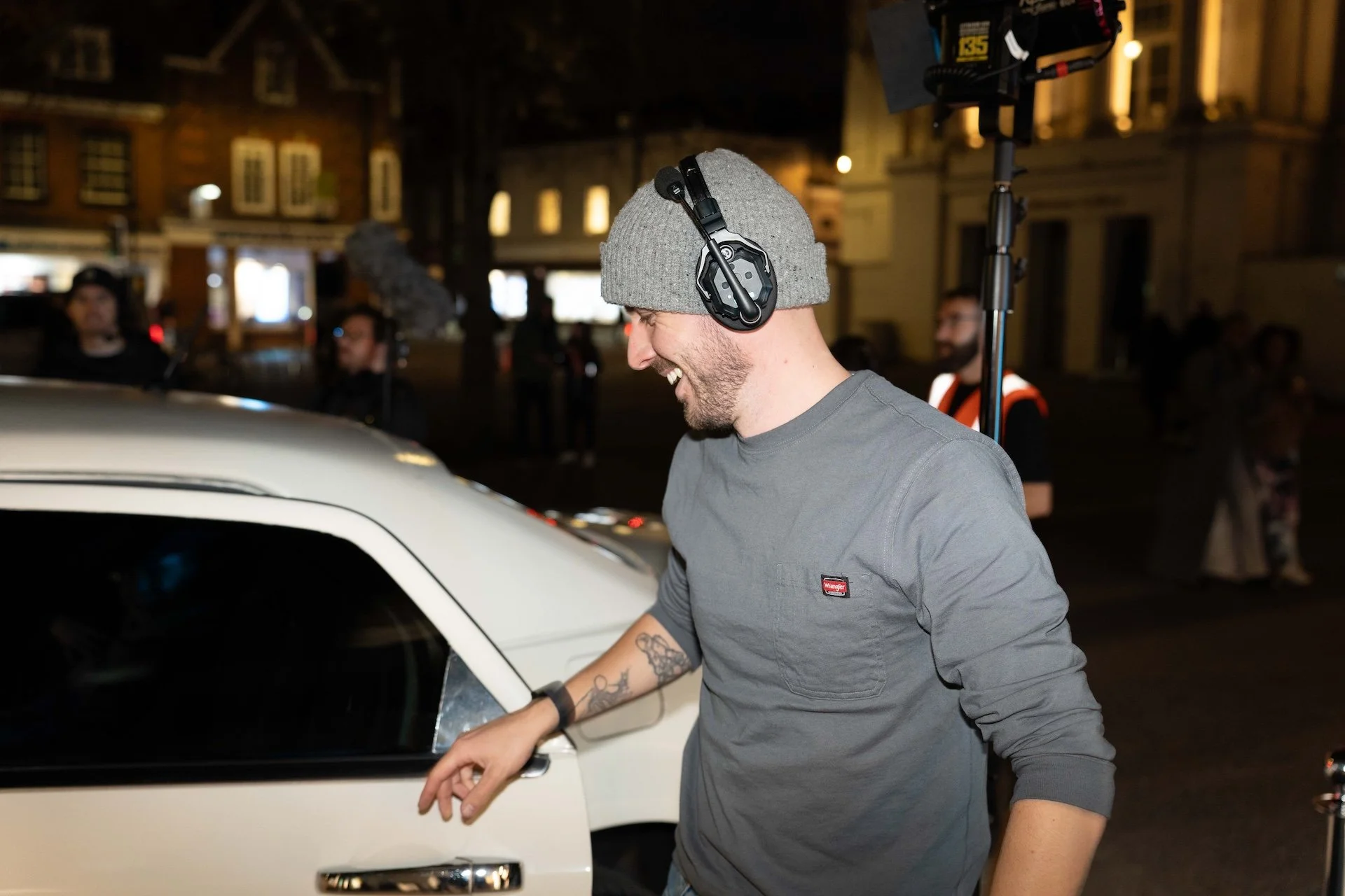 A young man wearing a gray beanie, black headphones, and a gray shirt with a small logo, smiles as he leans on a white car at night. There are people and buildings in the background.
