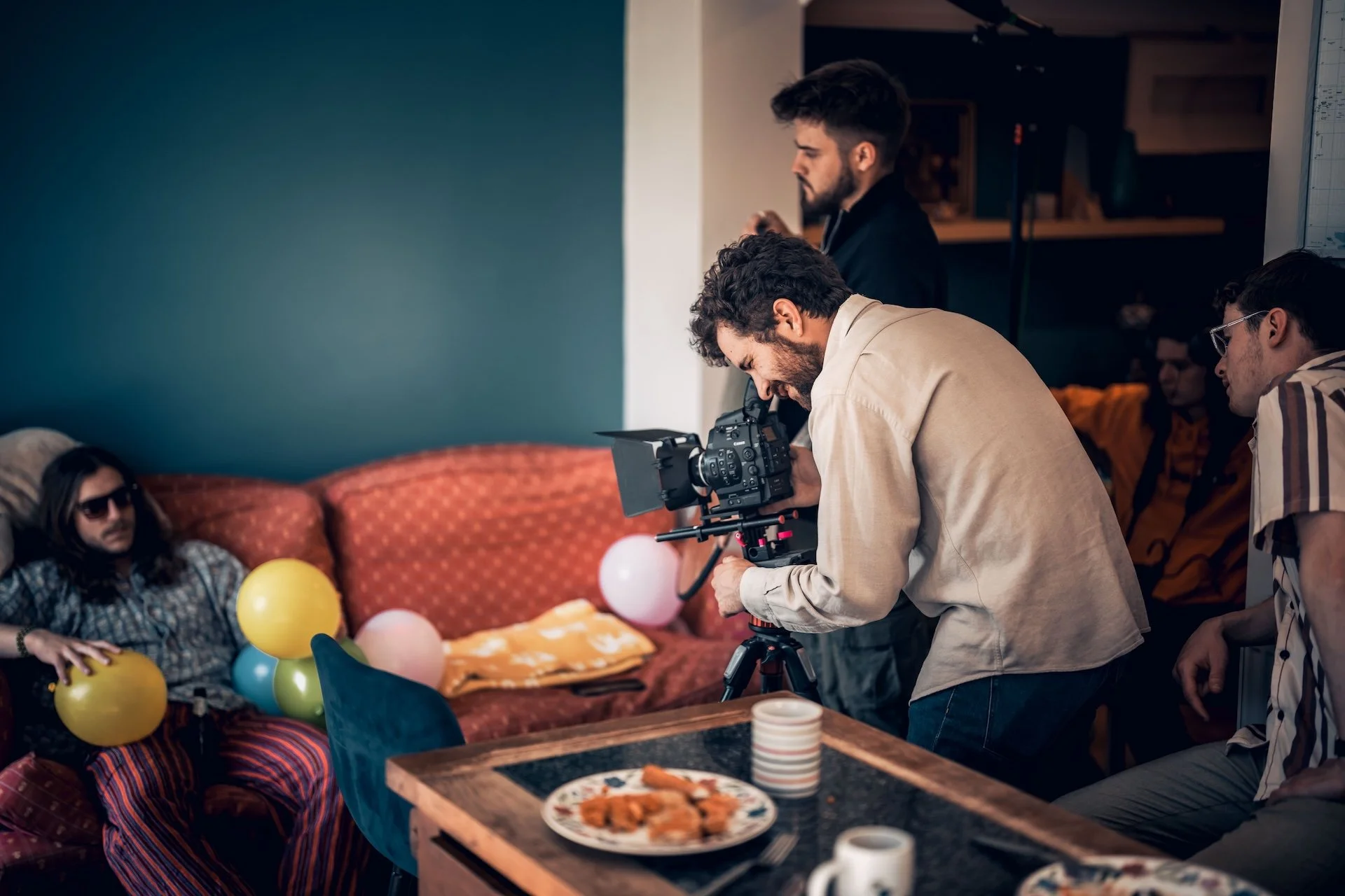 A group of people filming a man sitting on a sofa covered with colorful balloons during a party.