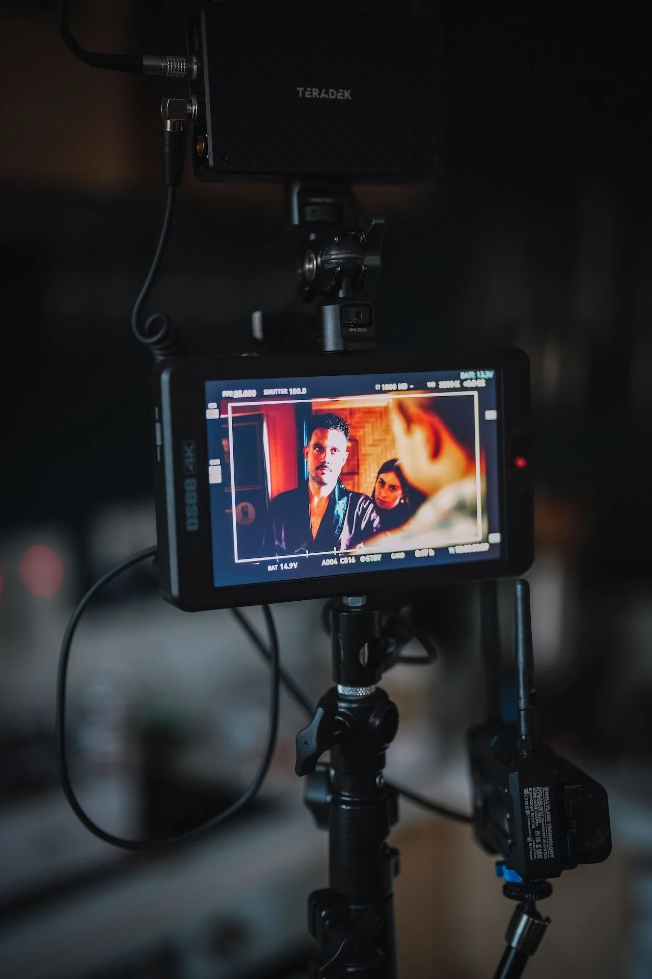 A camera monitor displays a scene from a film set with three actors engaged in conversation, with lighting and equipment visible in a dimly lit setting.