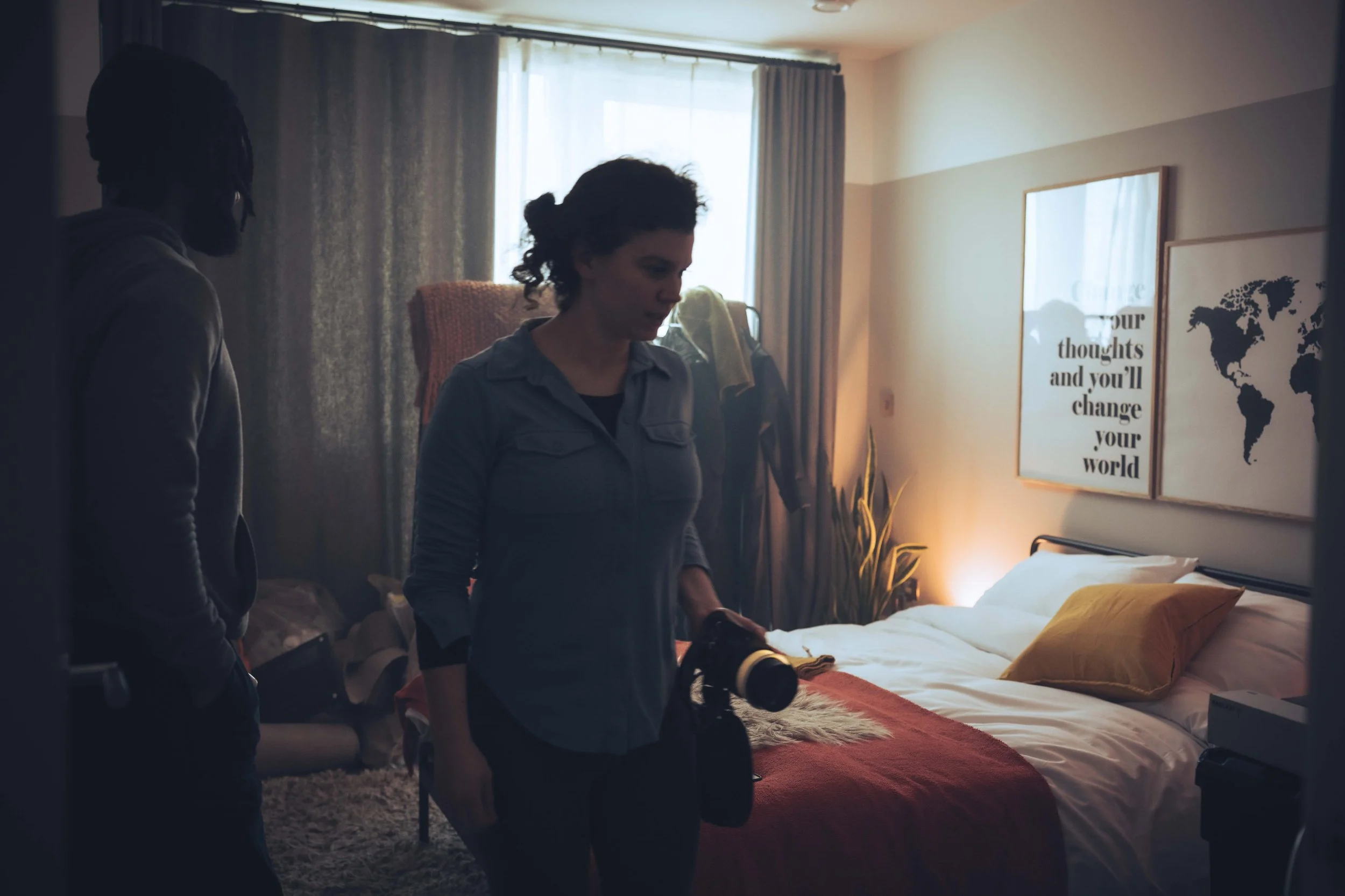 Two people standing in a bedroom, with sunlight coming through a window, beside a bed with yellow pillows and a red blanket. Two framed posters on the wall, one with a quote and the other with a world map.