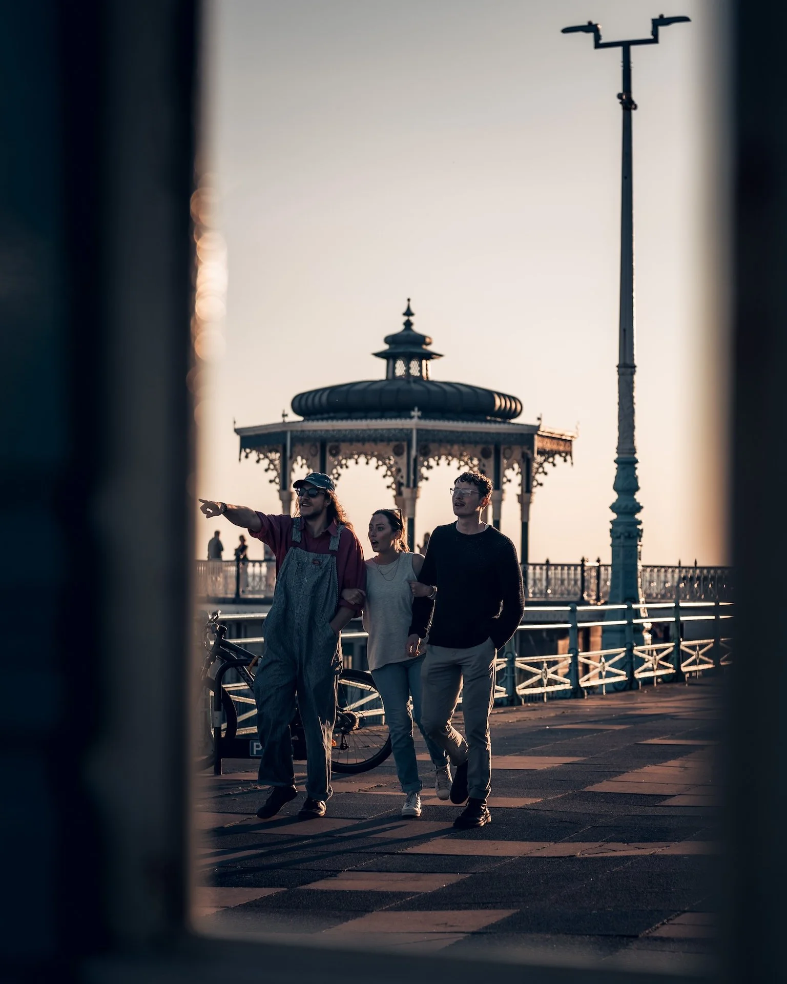 People walking along a pier with a gazebo and a lamppost during sunset.