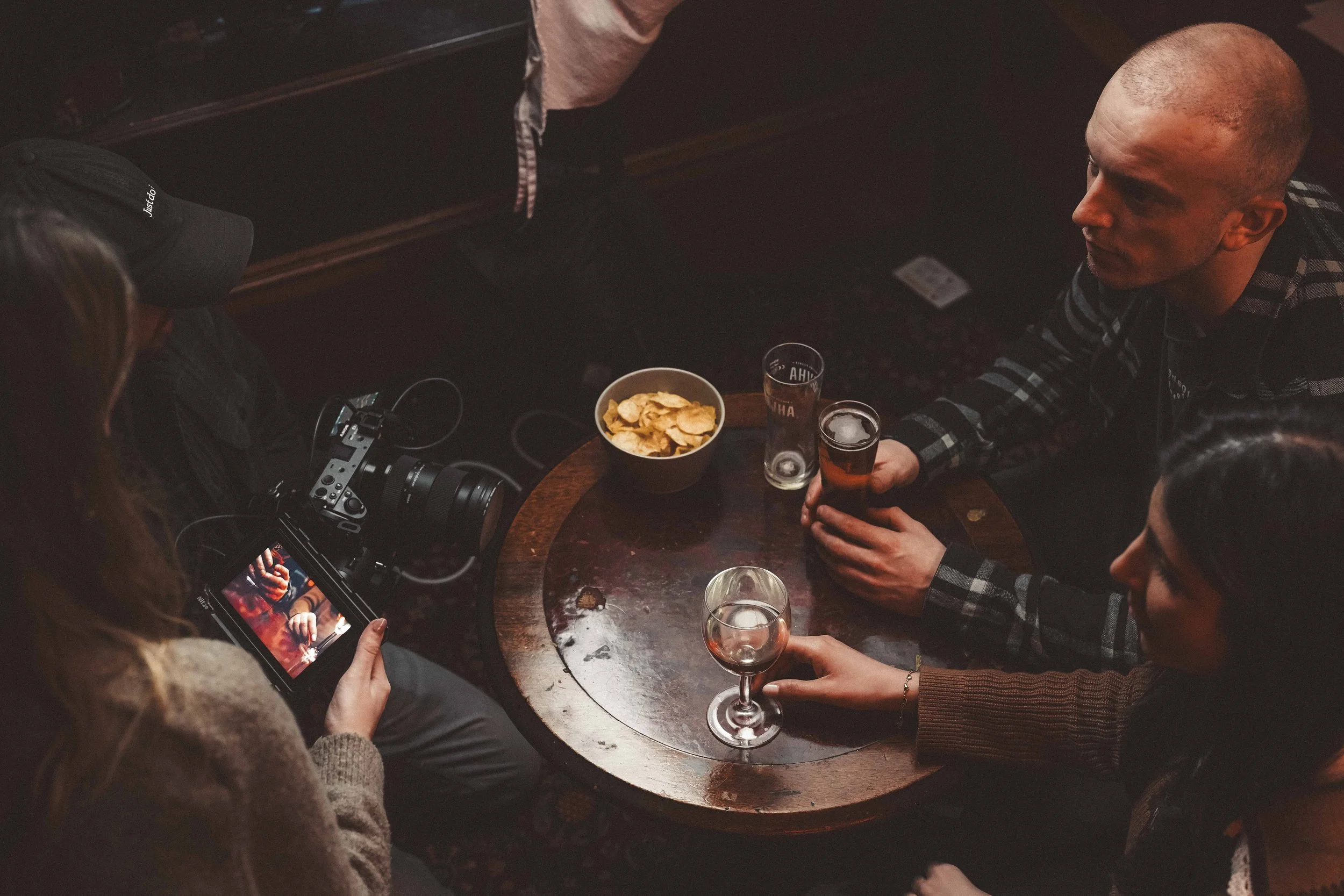 People gathered around a dark wooden table with drinks and snacks, including a bowl of potato chips, in a dimly lit setting. One person is holding a camera, taking a photo of the scene.