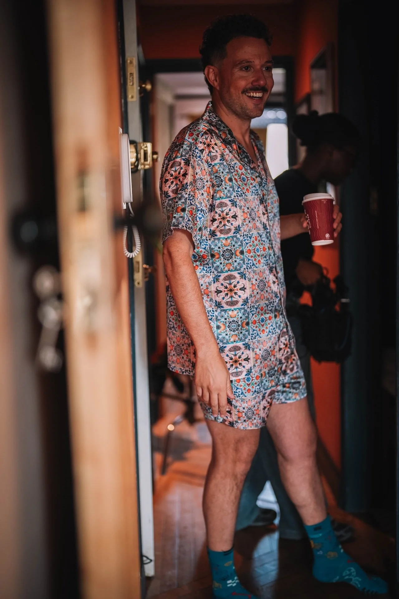 Man wearing a floral beach outfit, holding a red disposable coffee cup, smiling, standing in a hallway with dim lighting.