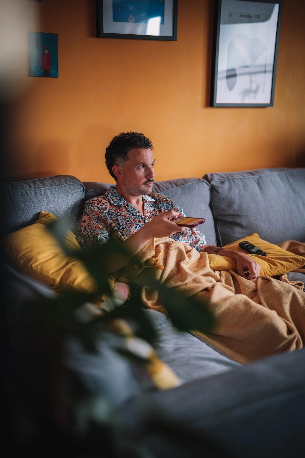 A man sitting on a couch with a yellow pillow, using a smartphone, with a TV on the orange wall behind him, and framed pictures on the wall. He is wrapped in a beige blanket, wearing a patterned shirt.