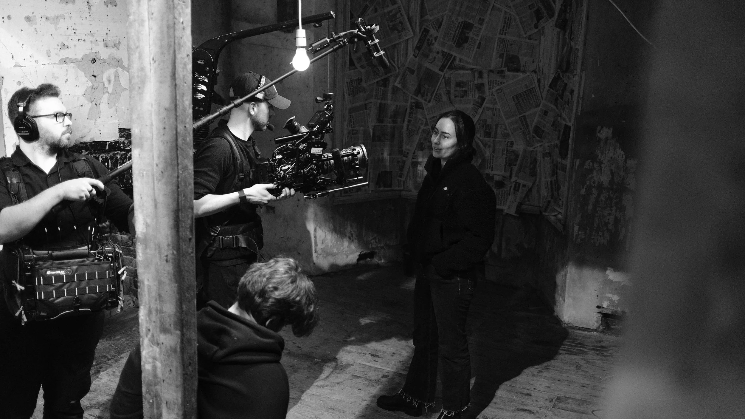 A woman being filmed on a video set with a camera operator and crew members in a room with newspaper-covered walls and distressed concrete.
