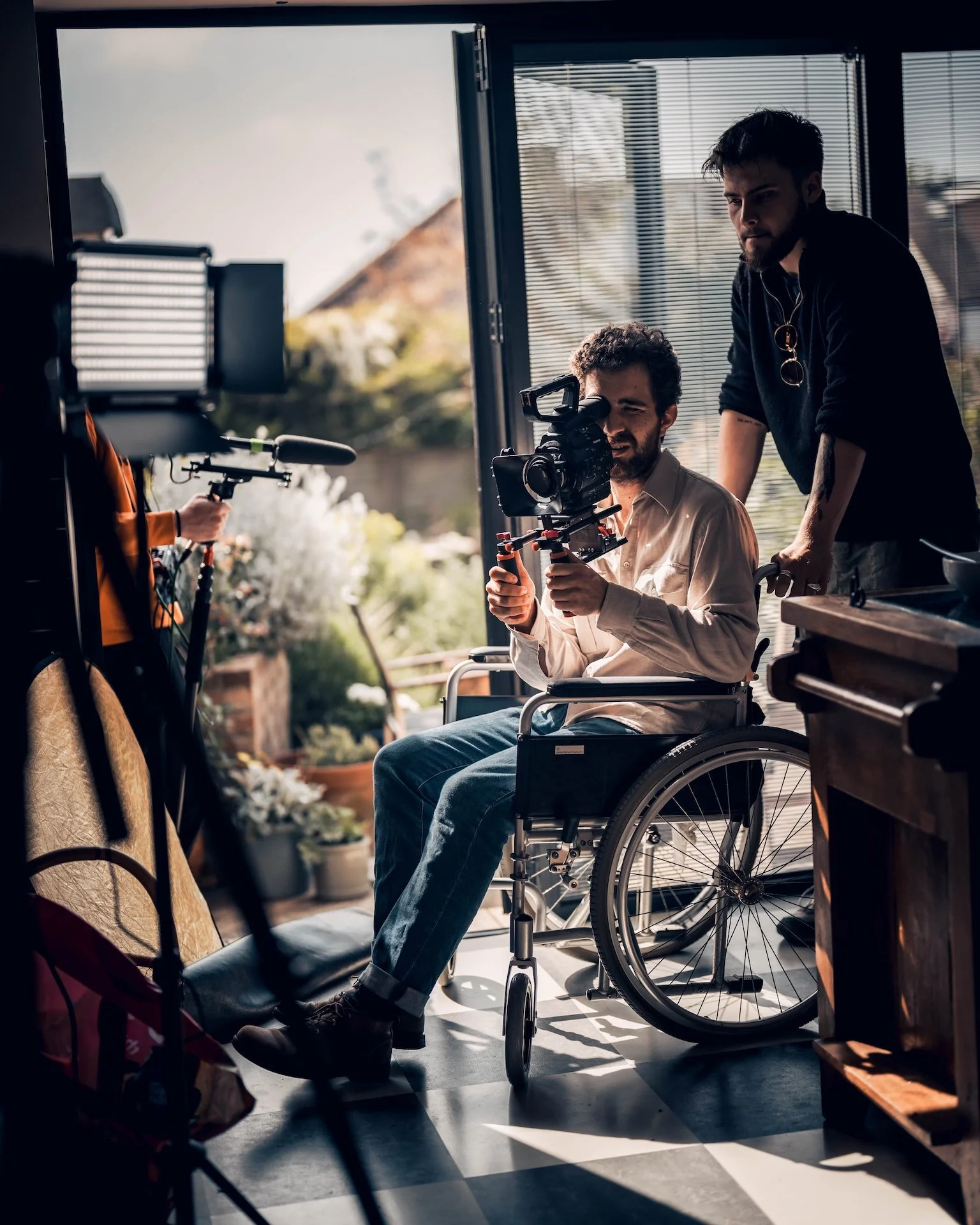 A man in a wheelchair filming a scene with a camera, another person holding a boom microphone, inside a room with sunlight coming through blinds, background showing a garden.