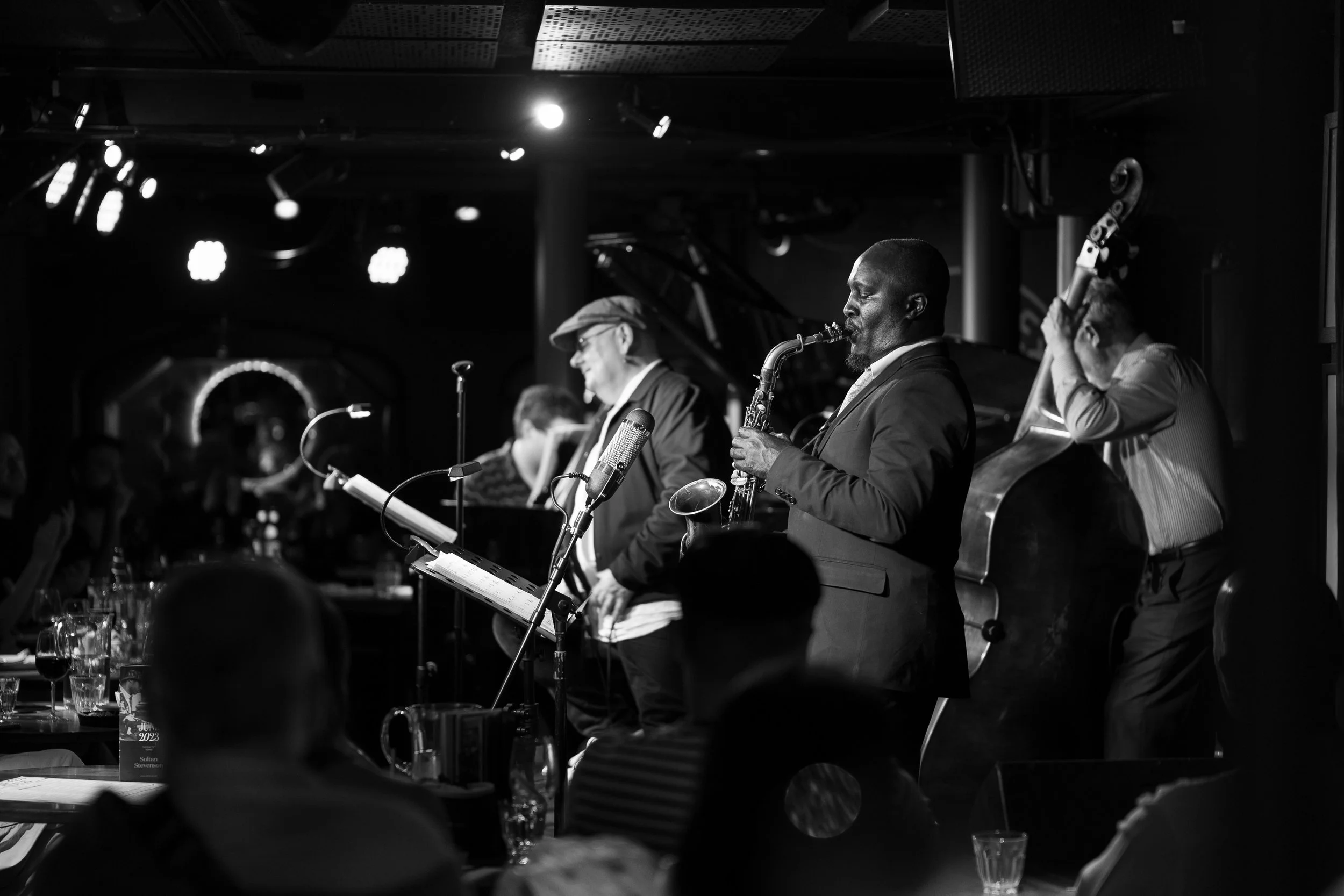 Jazz band performing on stage in a dimly lit venue, with audience members seated at tables in the foreground.