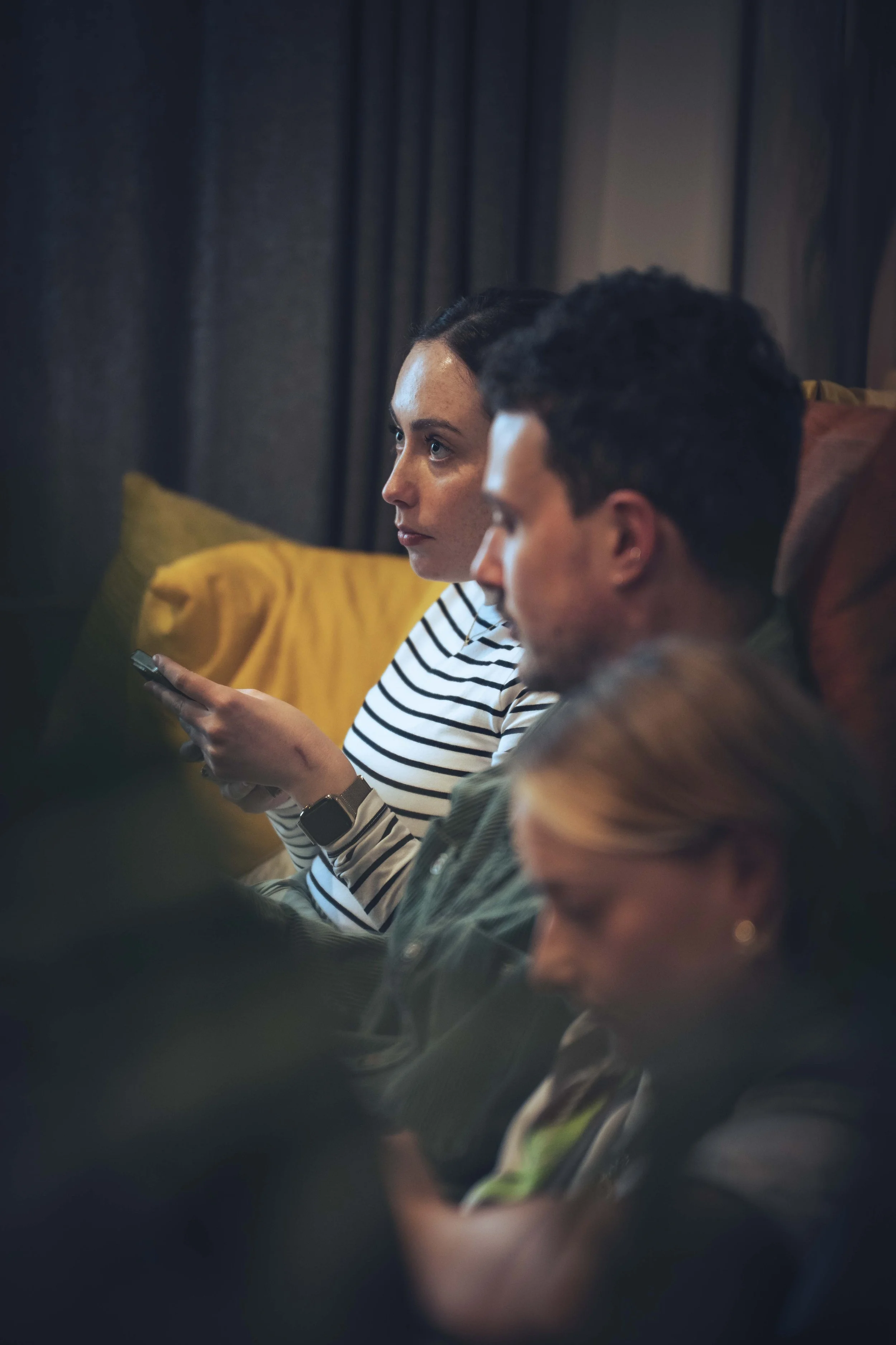 A woman with dark hair in a bun wearing a striped shirt and Smartwatch, and a man with dark curly hair in a brown jacket, both sitting on a couch in a dimly lit room with dark curtains and a yellow pillow in the background.