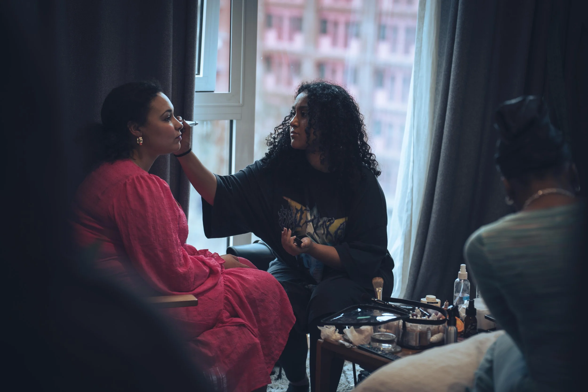 A makeup artist applying makeup to a woman seated by a window, with makeup products and brushes on a table nearby.