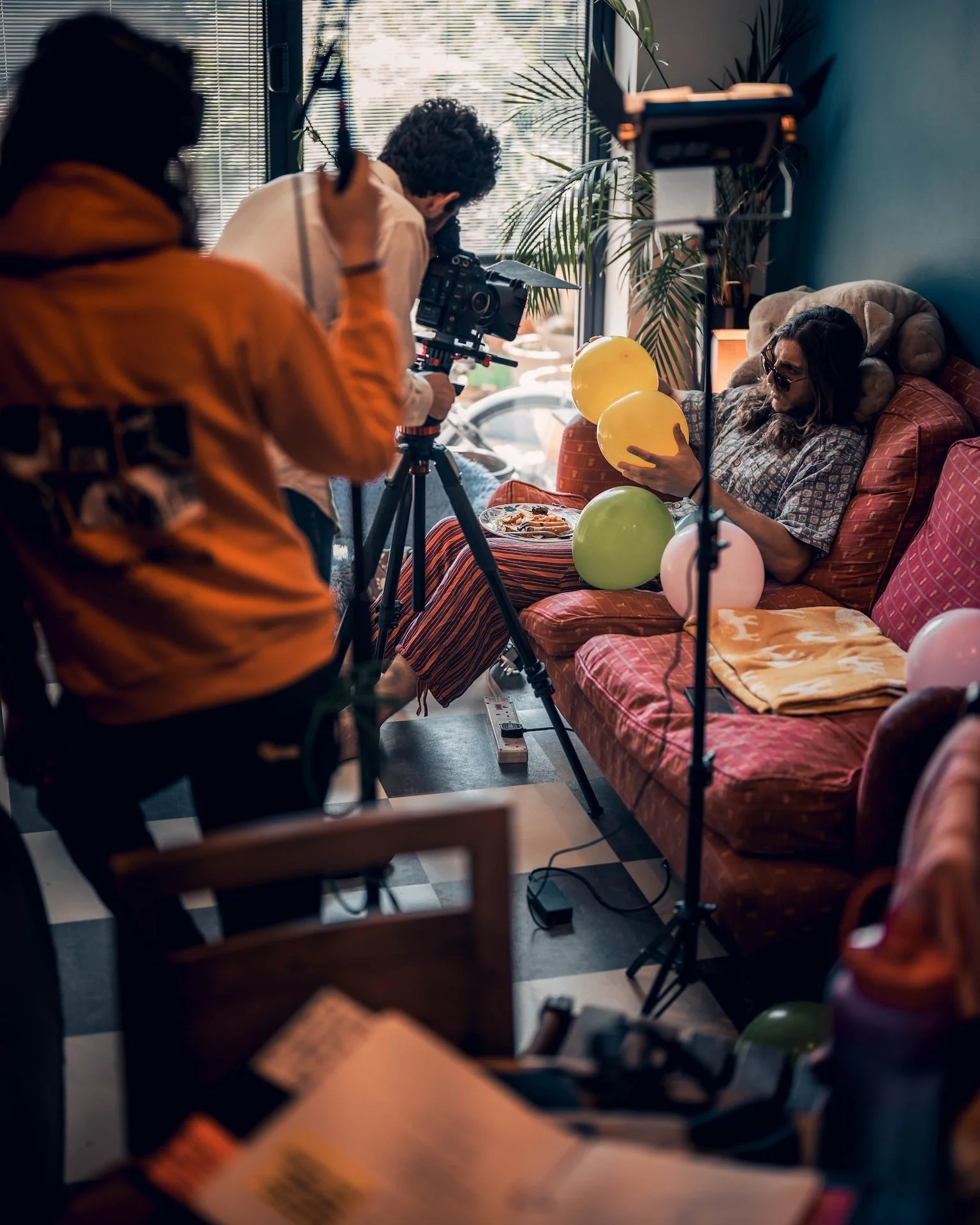 A person sitting on a red patterned sofa holding yellow and pink balloons, with a plate of food nearby, being filmed by a crew with a camera and lighting in a cozy room decorated with plants.