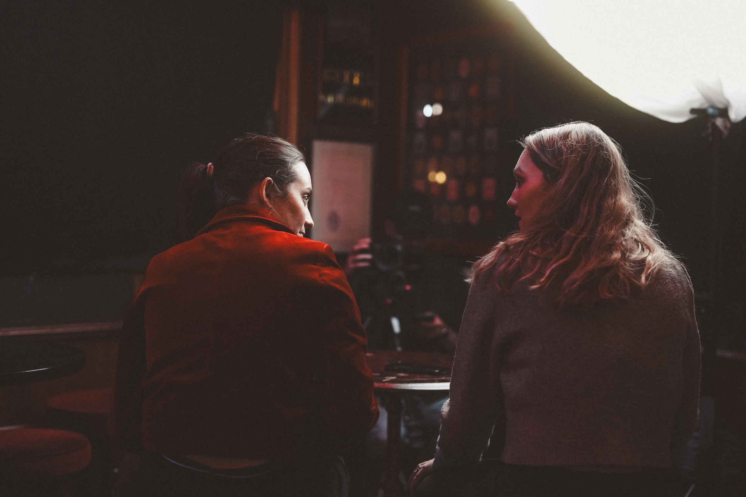 Two women with light skin sitting in a dimly lit room, facing each other and engaged in conversation. One woman has dark hair pulled back and is wearing a red jacket, while the other has curly blonde hair and is wearing a grey top. The background sho